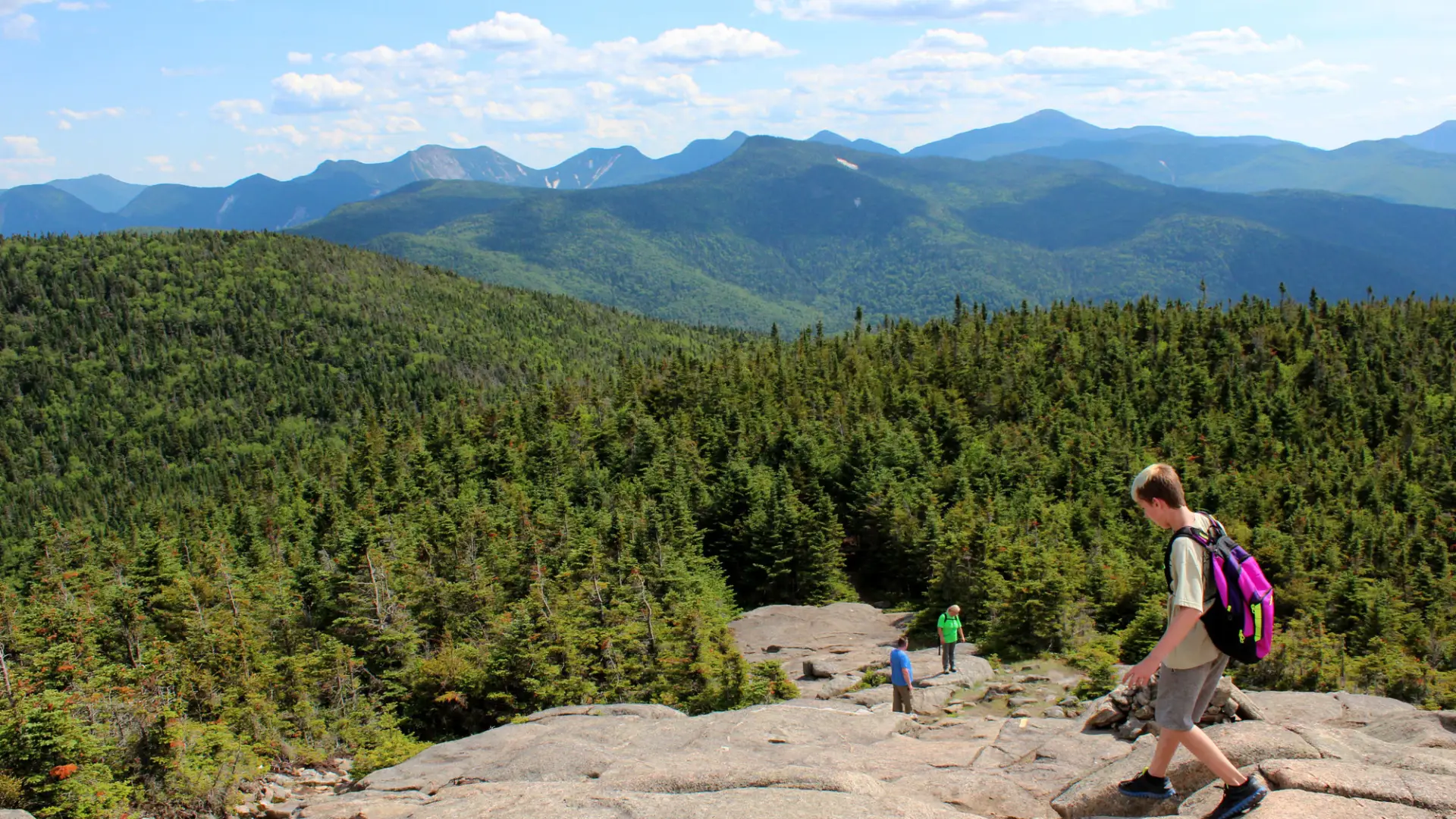 Views from the top of a hike with people hiking down a rock slab and mountain views in the background.