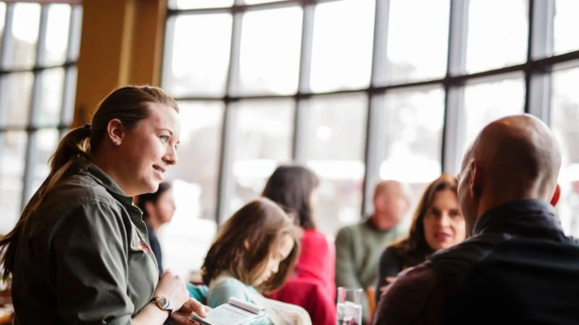 A waitress takes an order in front of a wall of windows.
