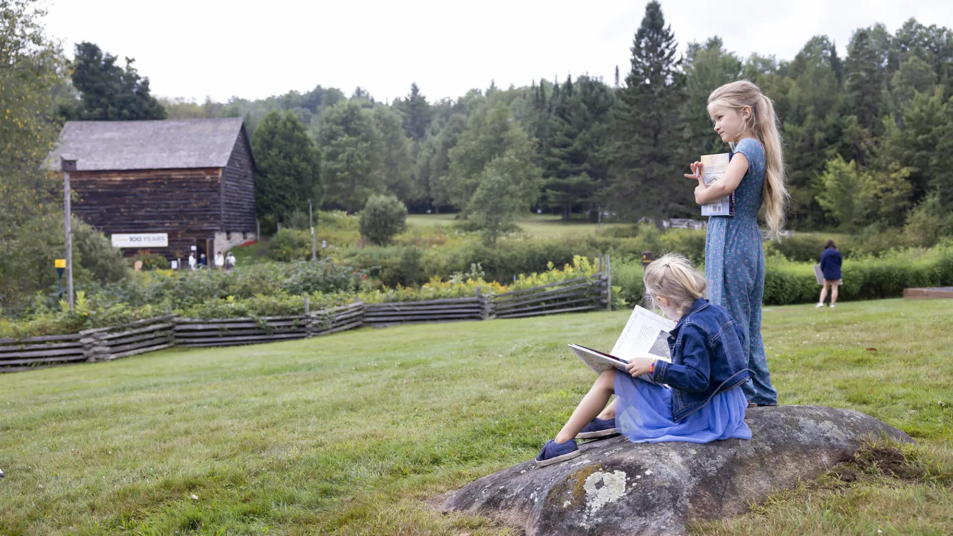 Girl sits on rock in dress and jacket reading book while second girl stands next to her holding book and looking out over green field with barn in distance