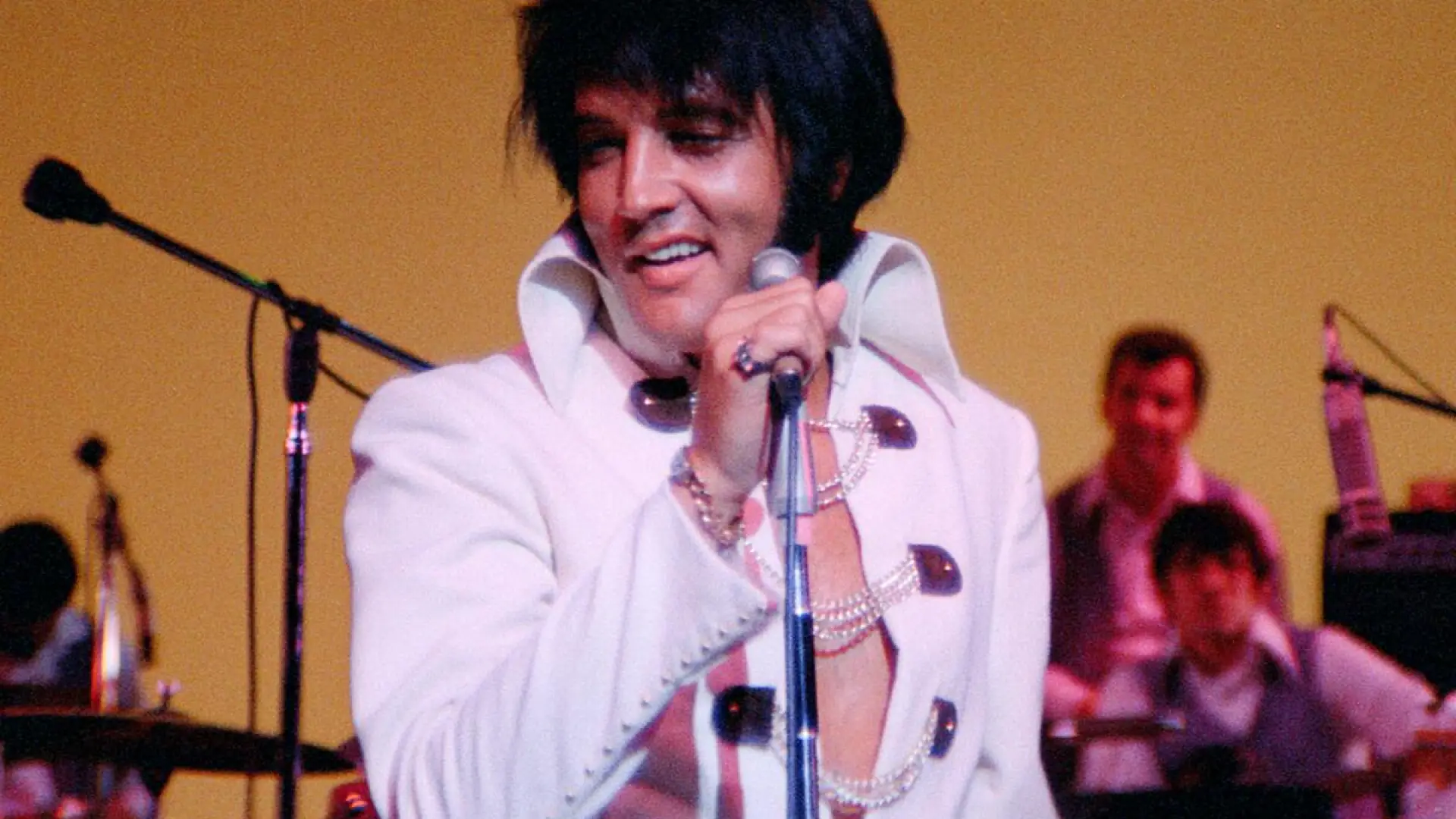 Still image of Elvis Presley in white costume on stage smiling behind microphone