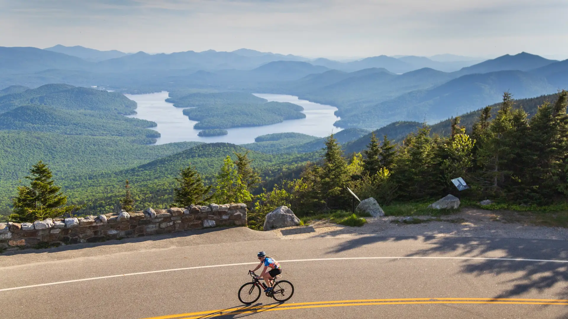 A cyclist rides along a mountain road overlooking a panorama of forested hills and an island-dotted lake in the valley below.