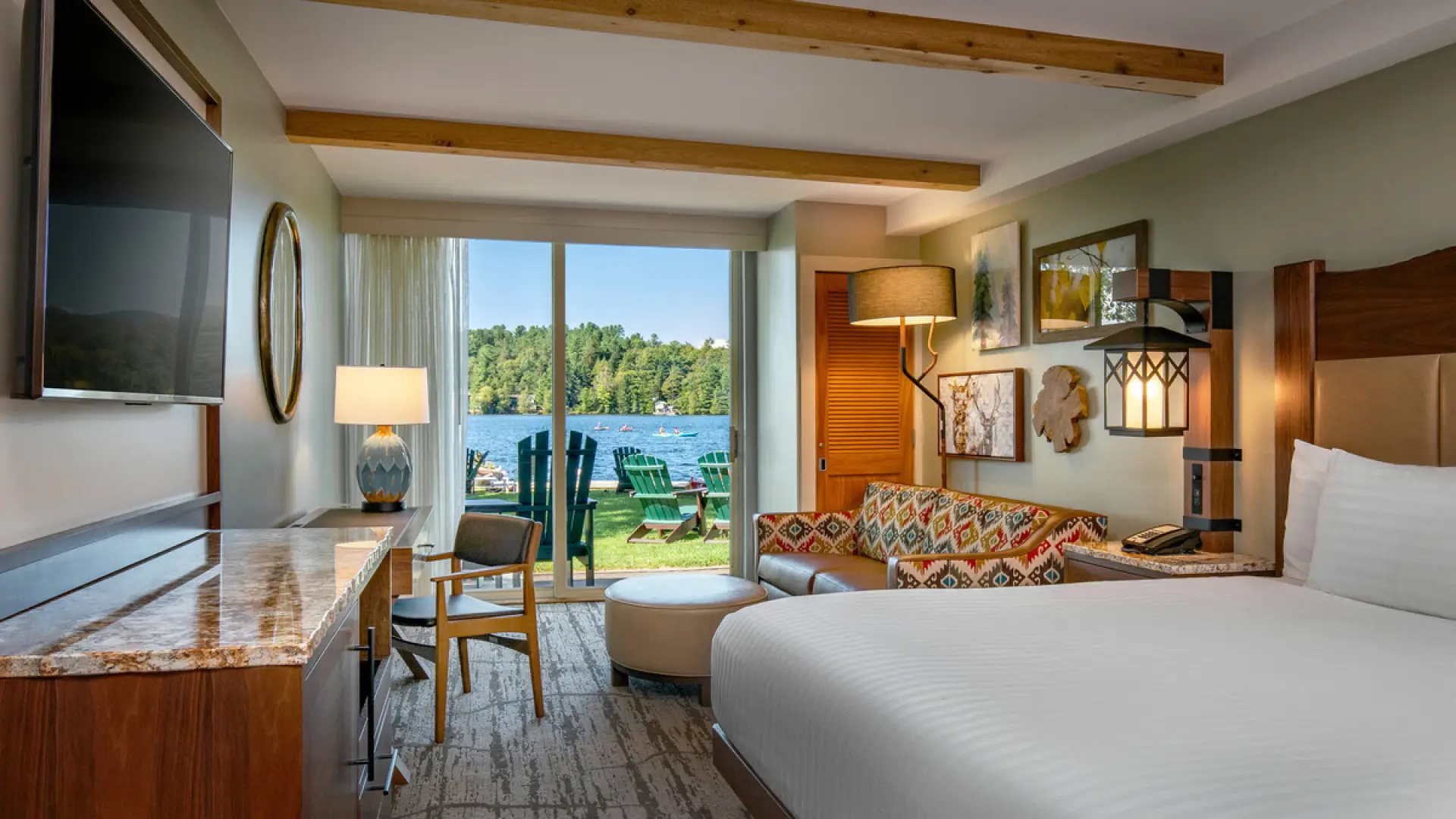 Hotel room with white bed and sitting area with sliding glass doors opening onto lakeside lawn with green Adirondack chairs