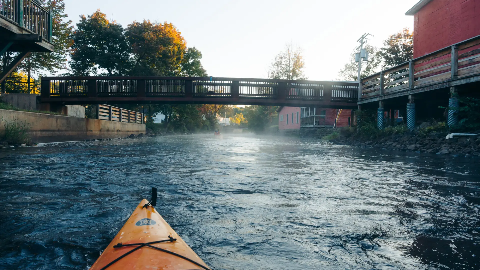 An orange kayak paddles down a misty river toward a low wooden bridge in the early morning light.