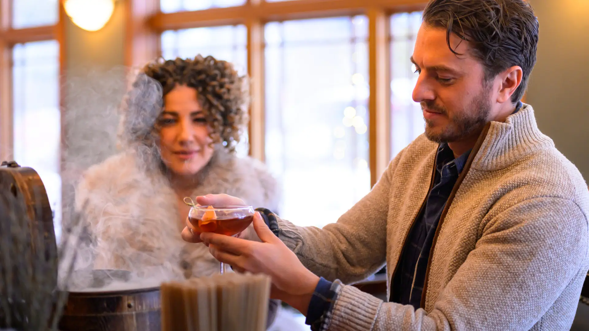 Man pulls cocktail from smoking box at bar as woman looks on