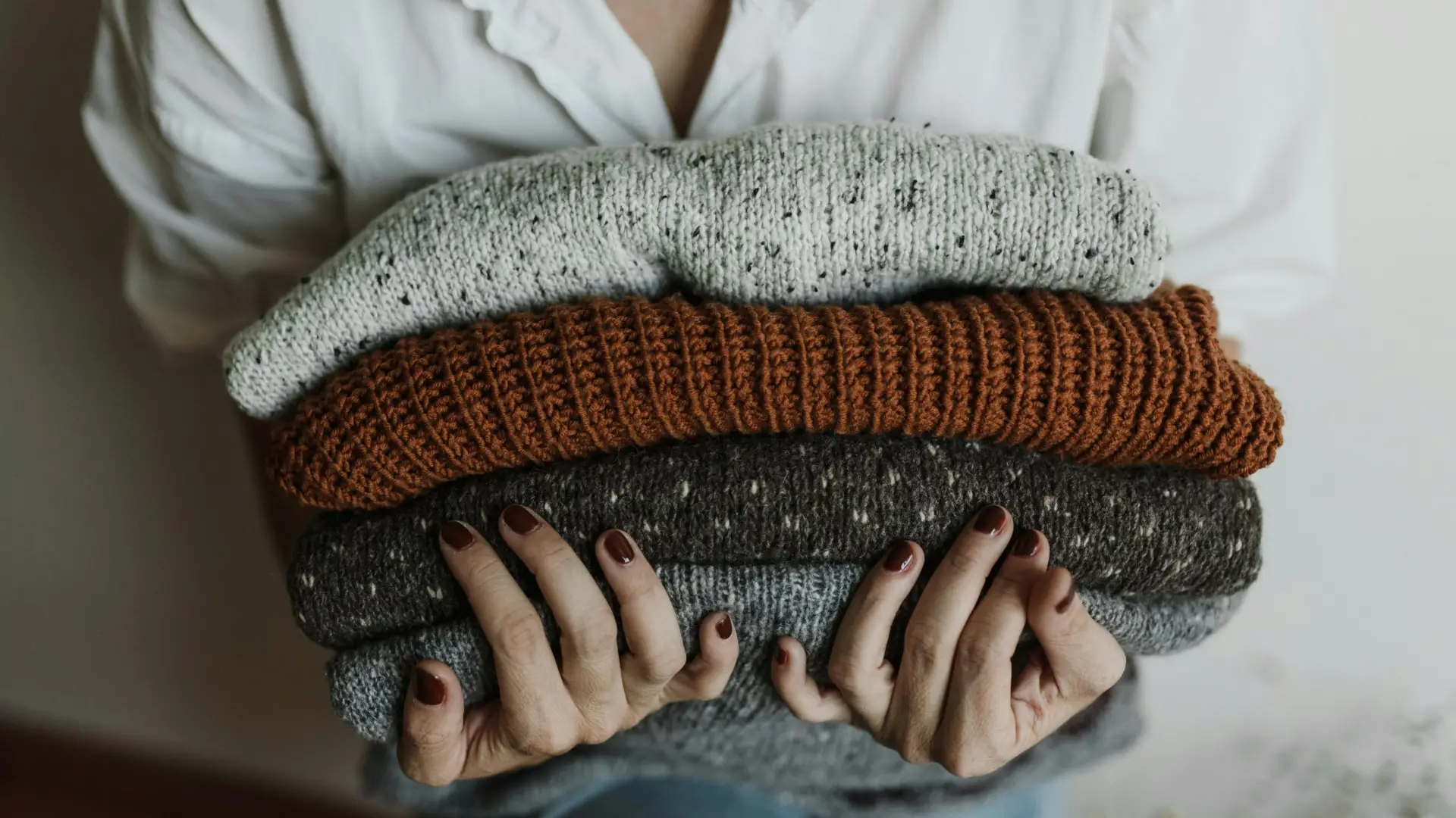 Person wearing white dress shirt and with nails painted a deep orange holds a stack of knit sweaters in her arms