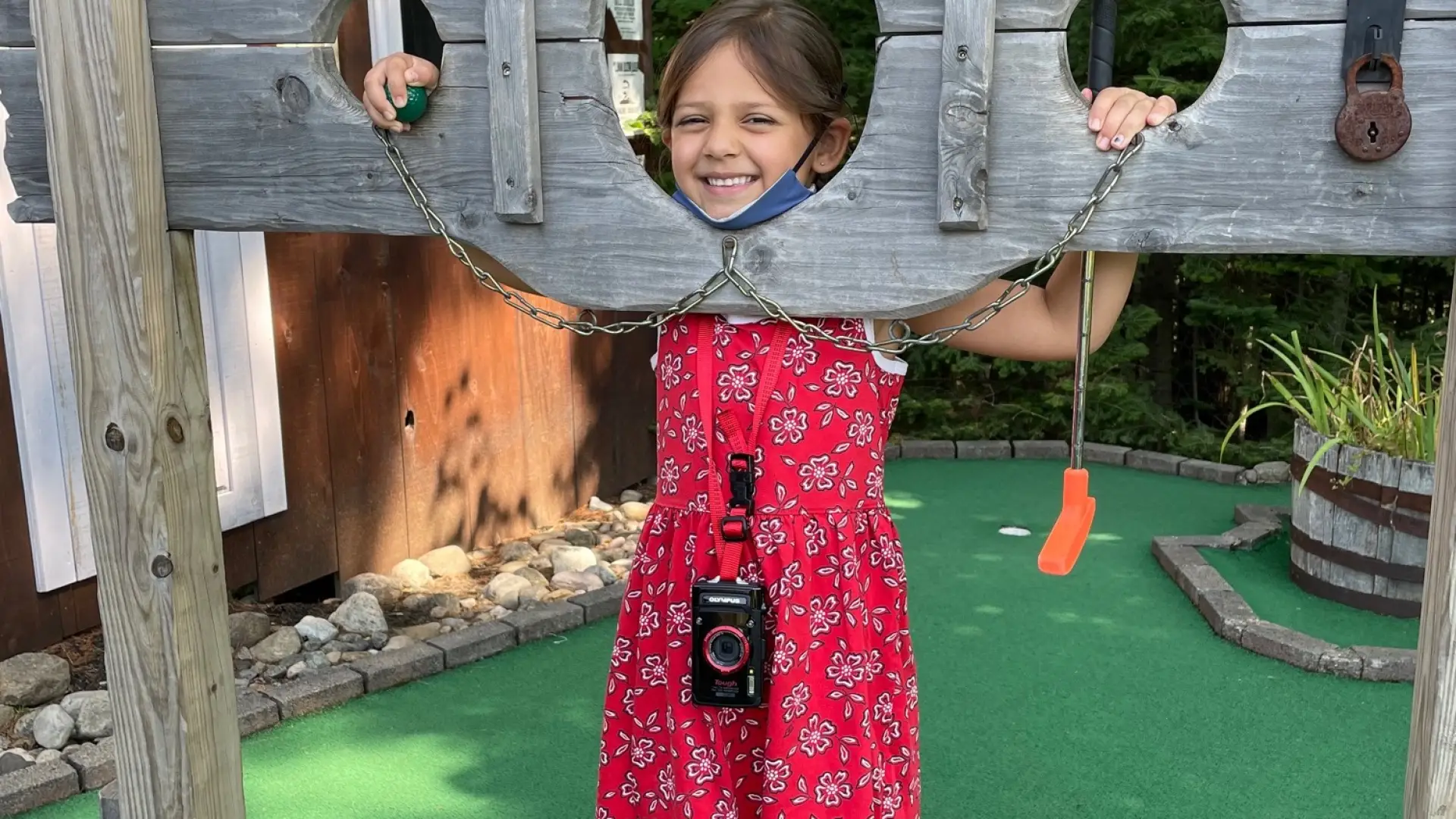 A small girl in a red dress poses in a wooden pillory on a mini golf course