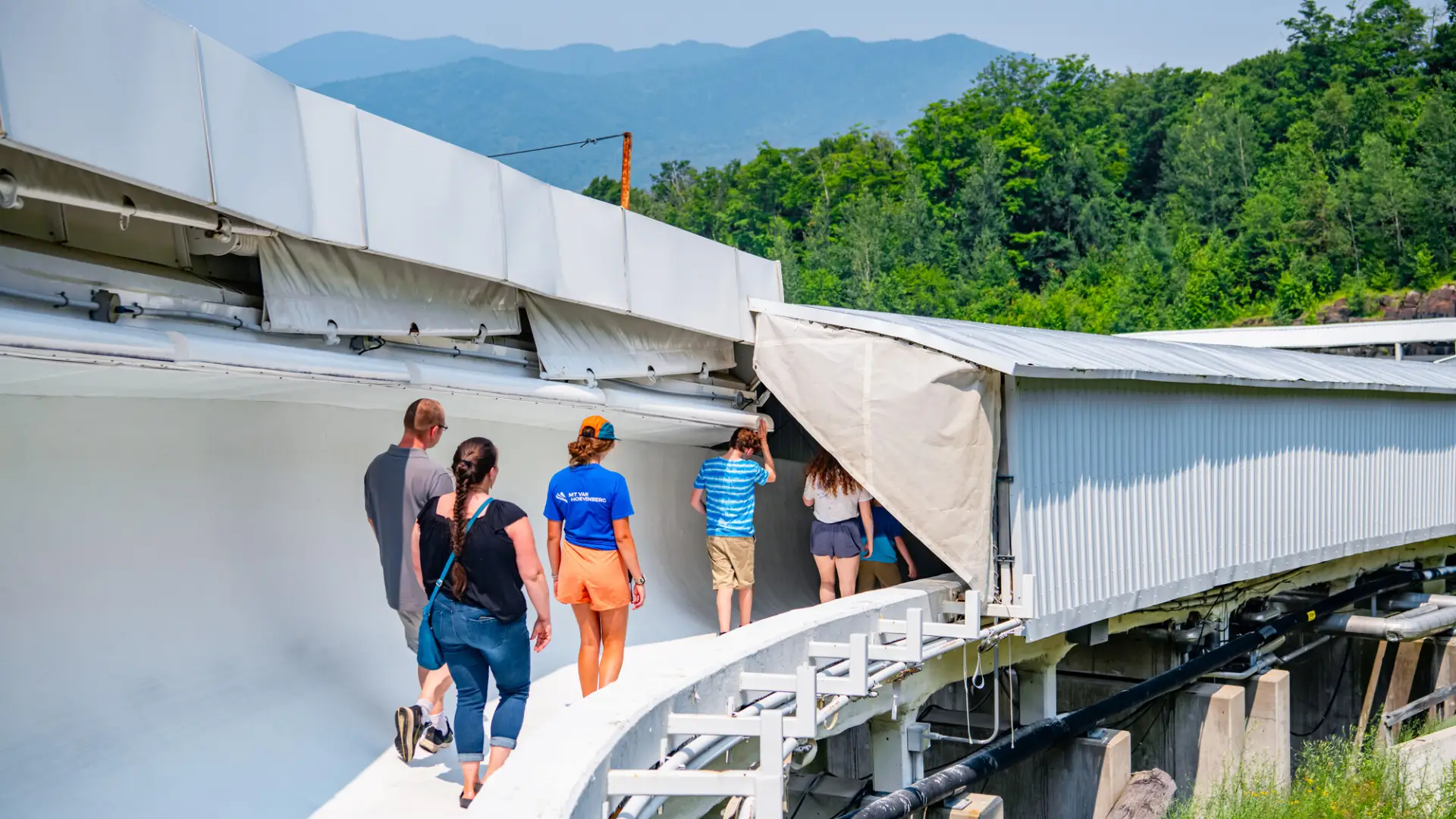 Family of five and female tour guide walk on bobsled track in summer with mountain views in background