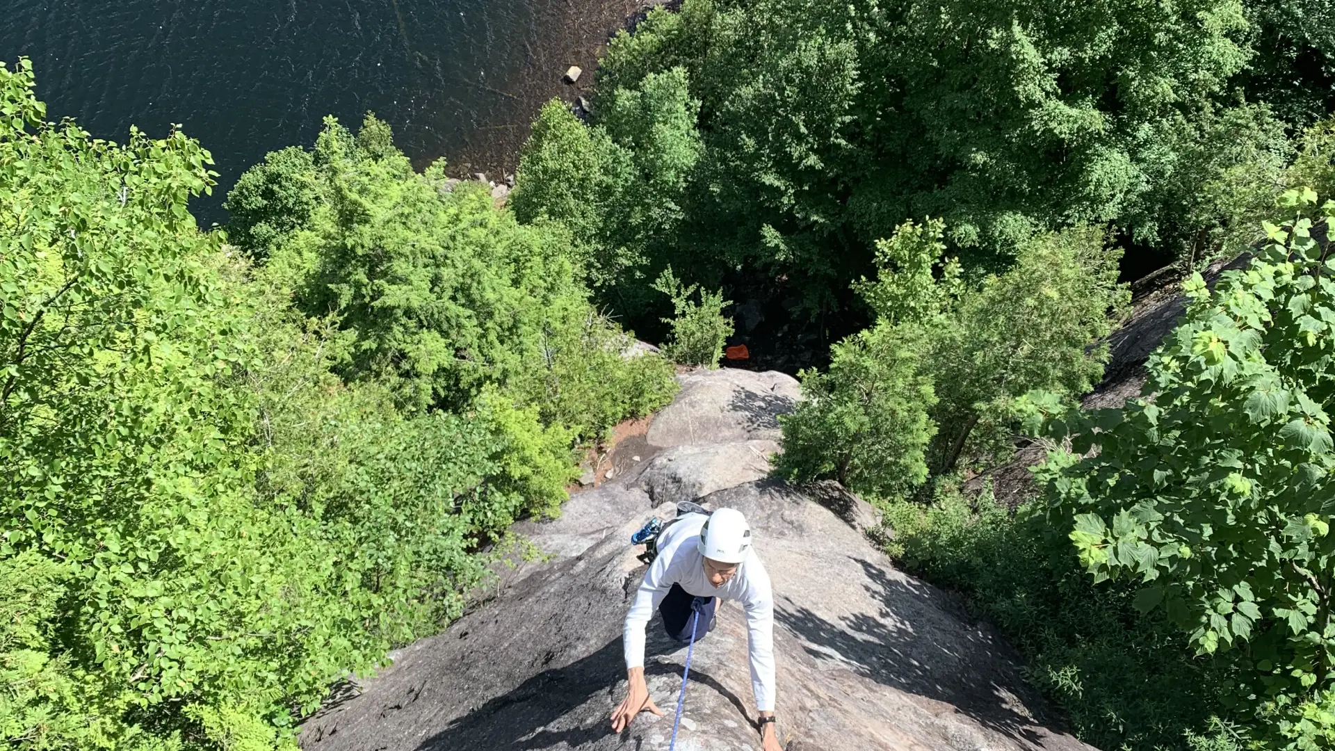 A rock climber ascends a steep cliff face overlooking a serene lake nestled within a lush green forest.