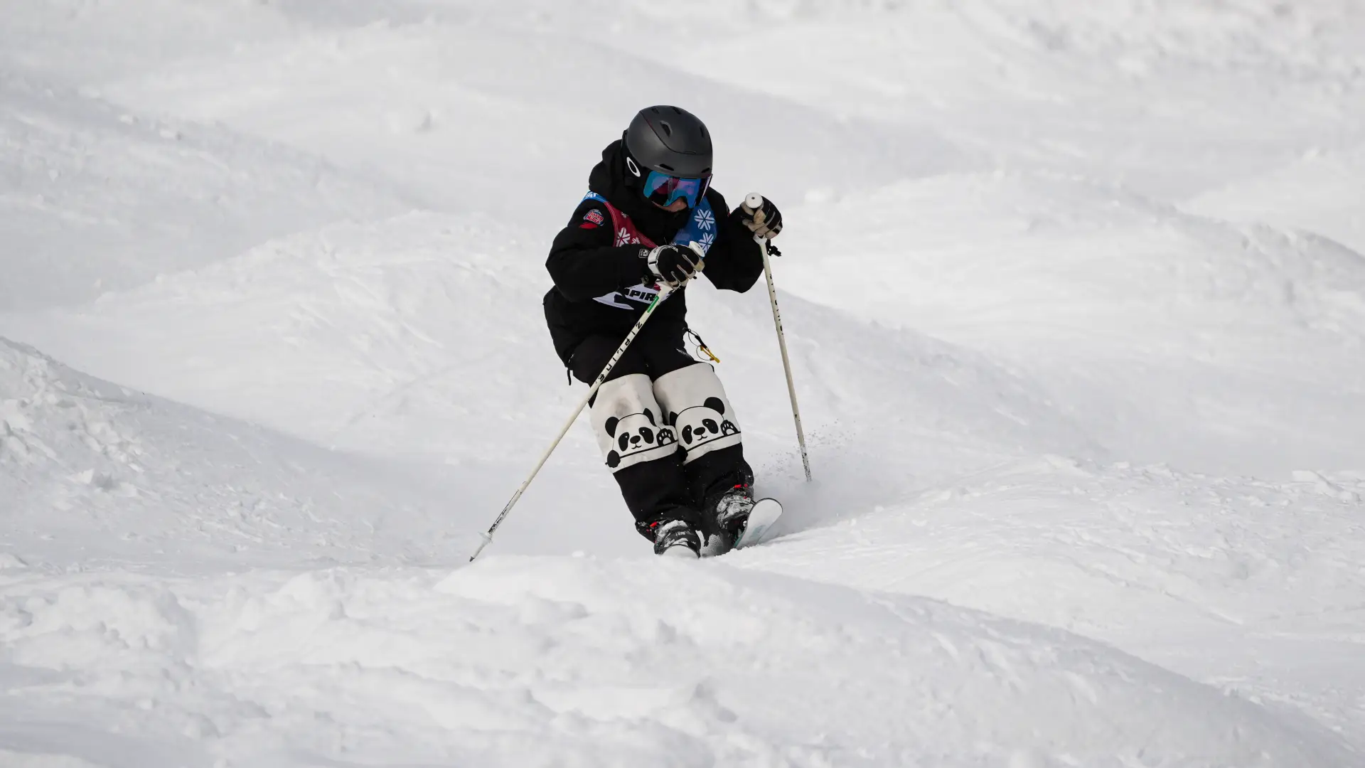A skier wearing black gear and panda-patterned knee pads navigates a mogul field on a snowy slope.