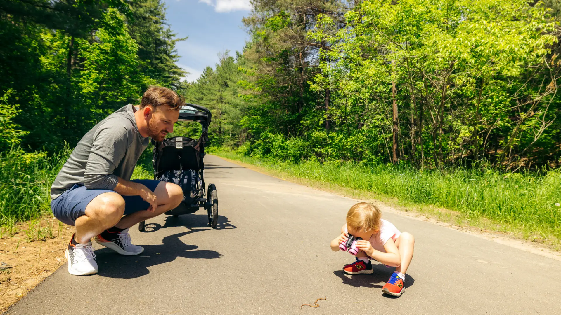 A man watches as his toddler studies an earth worm on a paved path.