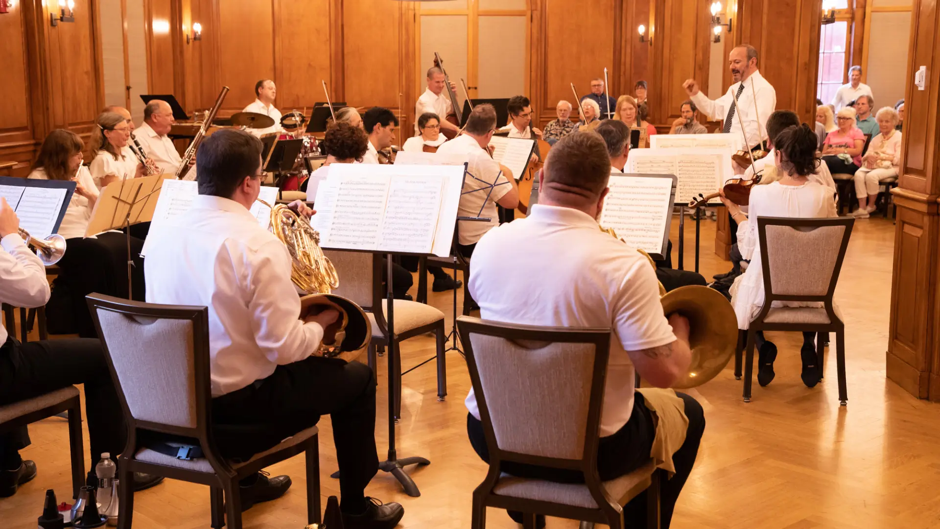 Lake Placid Sinfonietta performs under the direction of a conductor in a wood-paneled hall filled with audience members.