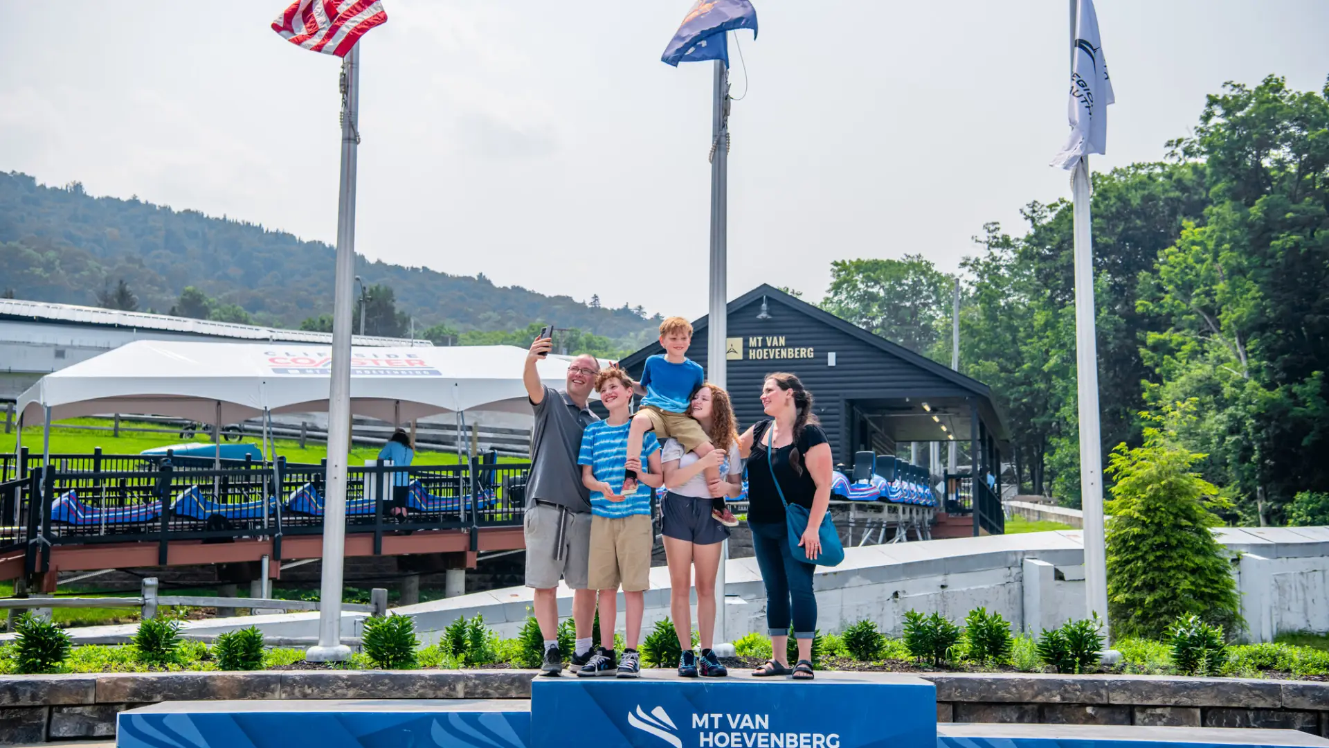 Mother, father, daughter, and older son stand with youngest son on their shoulders taking a selfie on 1st place podium at Mt Van Hoevenberg sliding complex