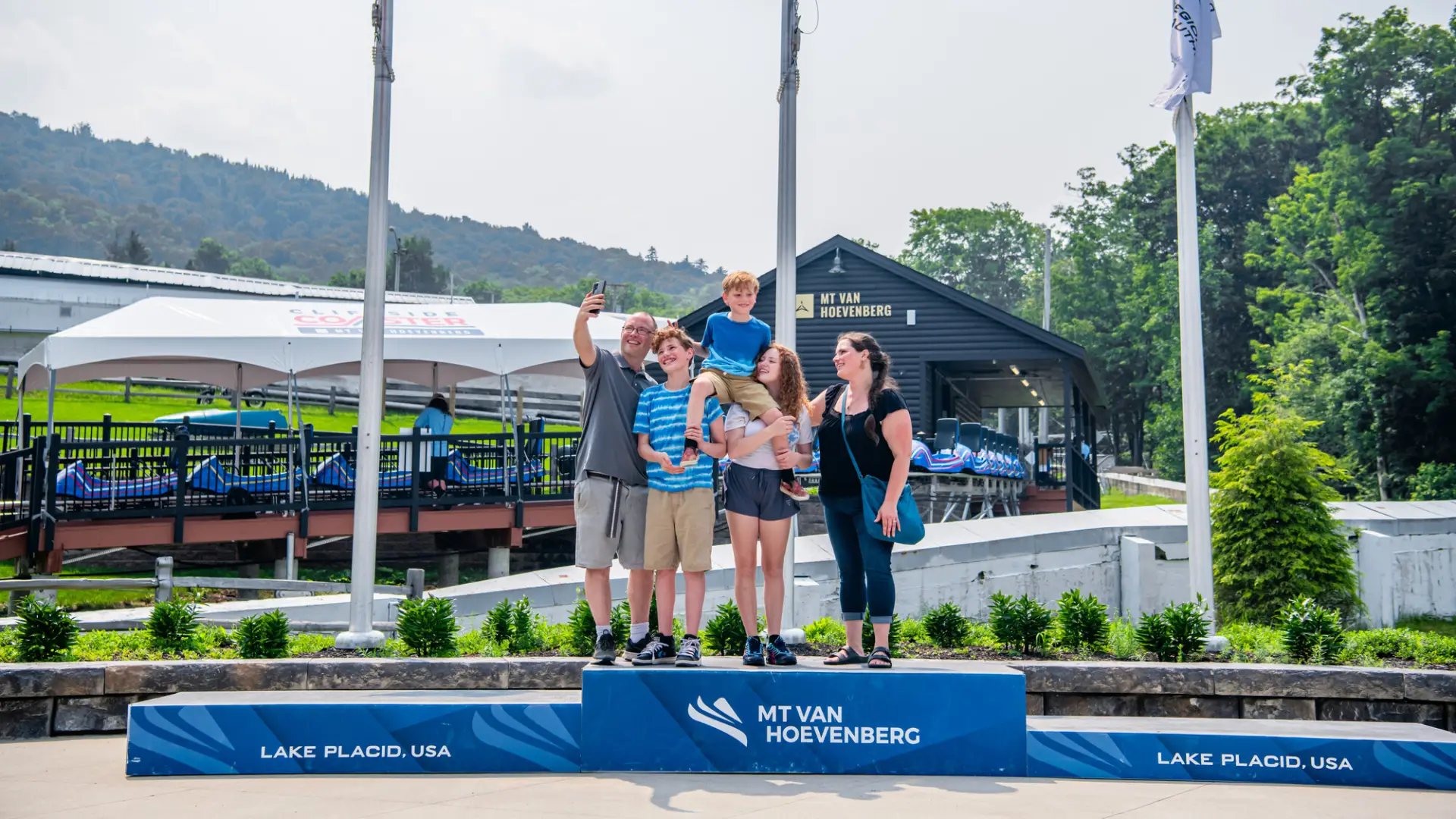Family of five stands on first place block of podium holding youngest child aloft and taking a selfie