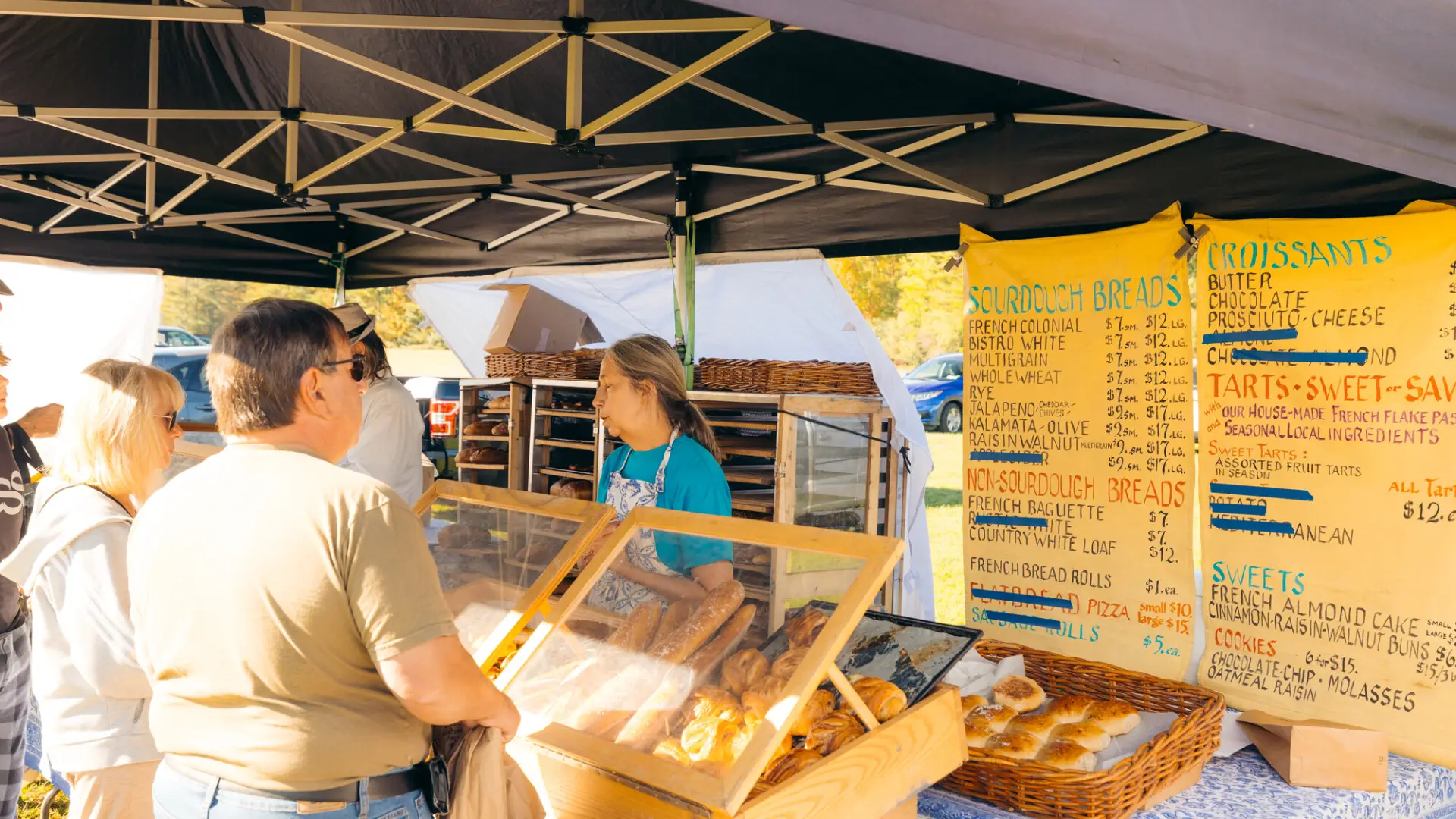 Person talks to seller over a table full of baked goods under outdoor farmers market tent