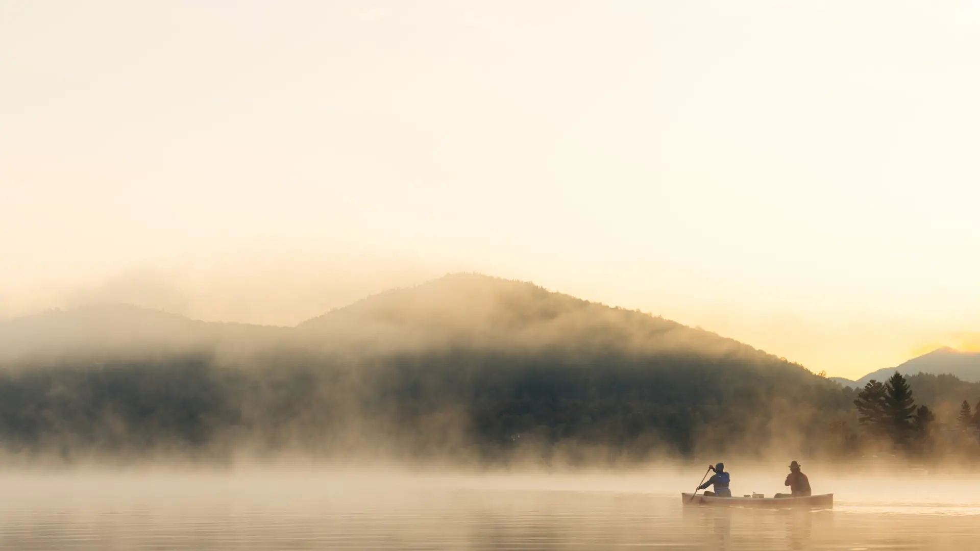 A canoe with two paddlers floats on mist-covered lake with mountain in background