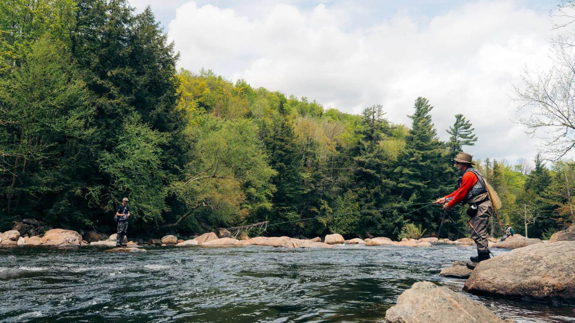 Two fly fishermen in waders and vests stand on large river rocks casting their lines into a wide, flowing stream.