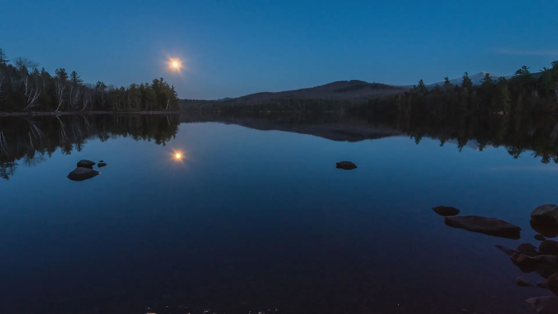 A tranquil lake at twilight reflects a full moon and the dark silhouettes of trees and mountains.  Several dark rocks are visible near the shore.