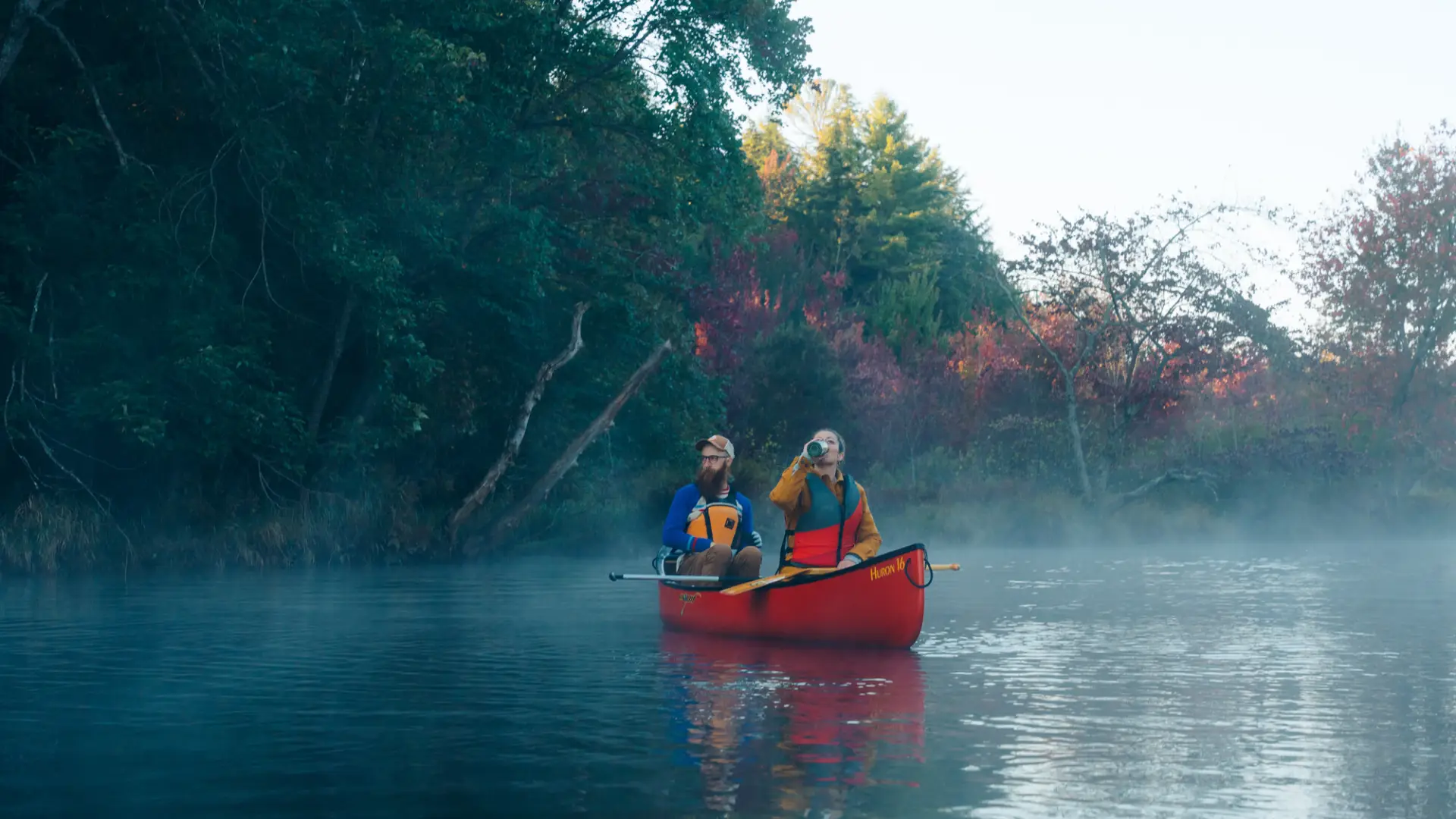 Man paddles at back of red canoe on foggy river while woman in front drinks from green thermos, both wearing life vests