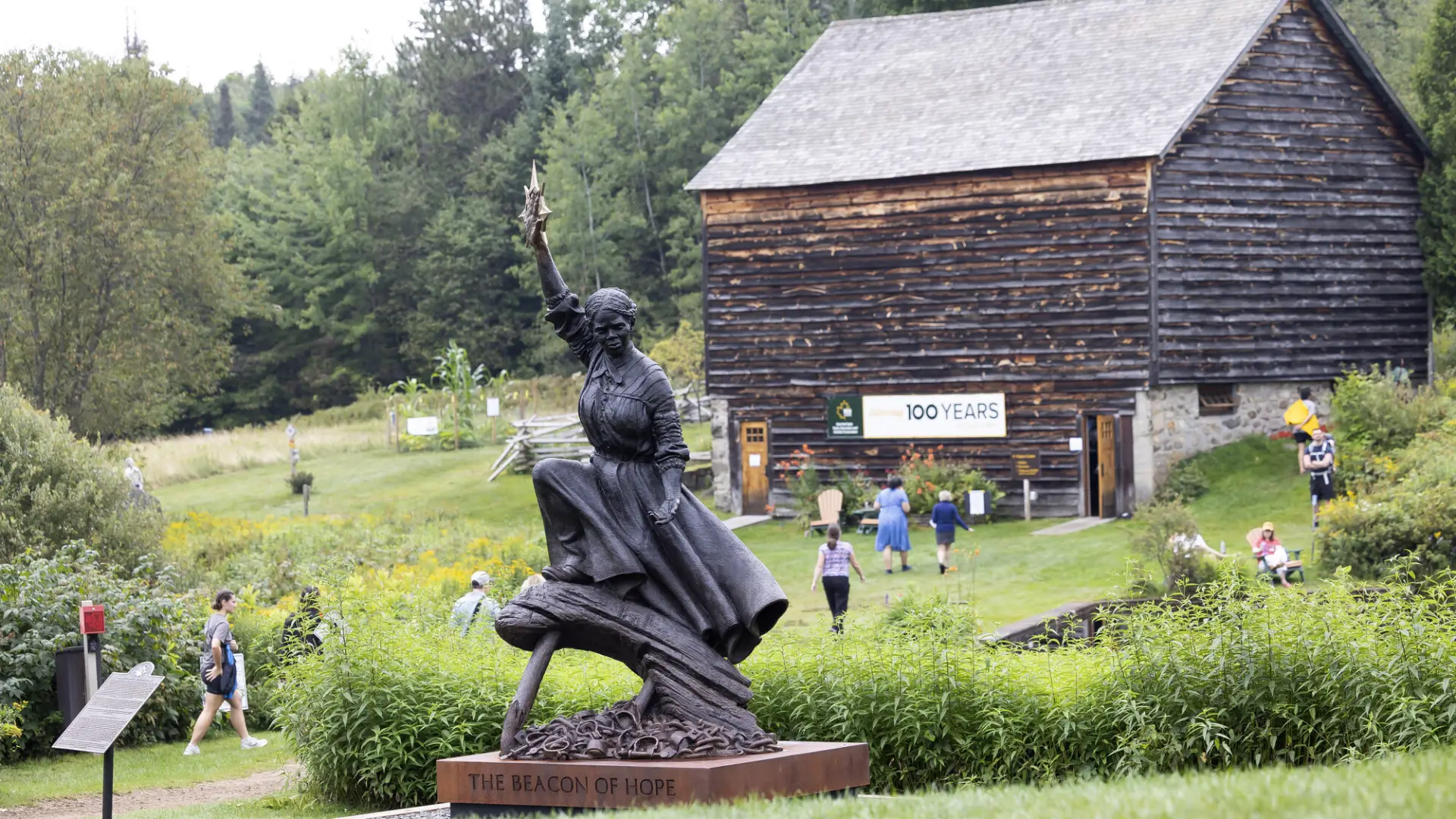 Harriet Tubman statue placed on green lawn with old barn in background and people walking on grounds