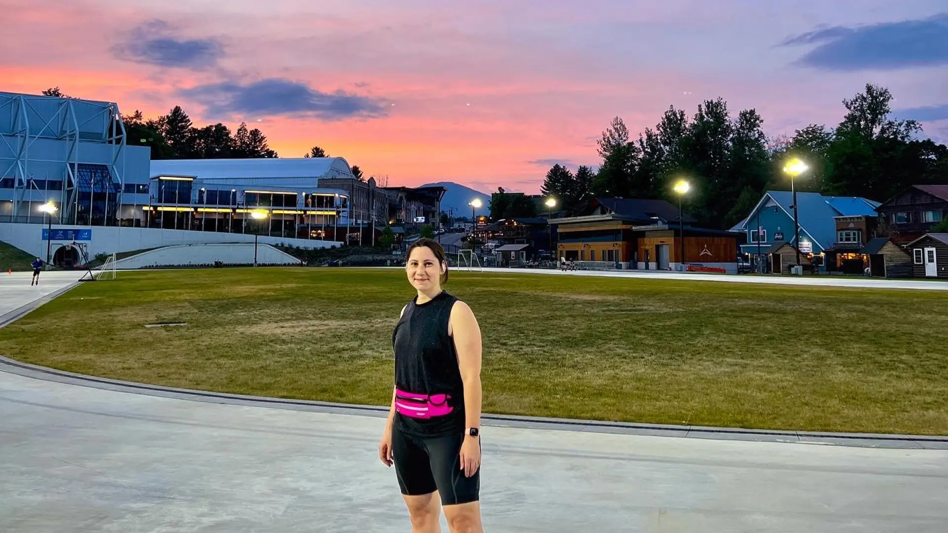 A woman stands on a skating oval wearing black and pink skating clothes and inline skates, while the sun is pink with a sunset behind her.