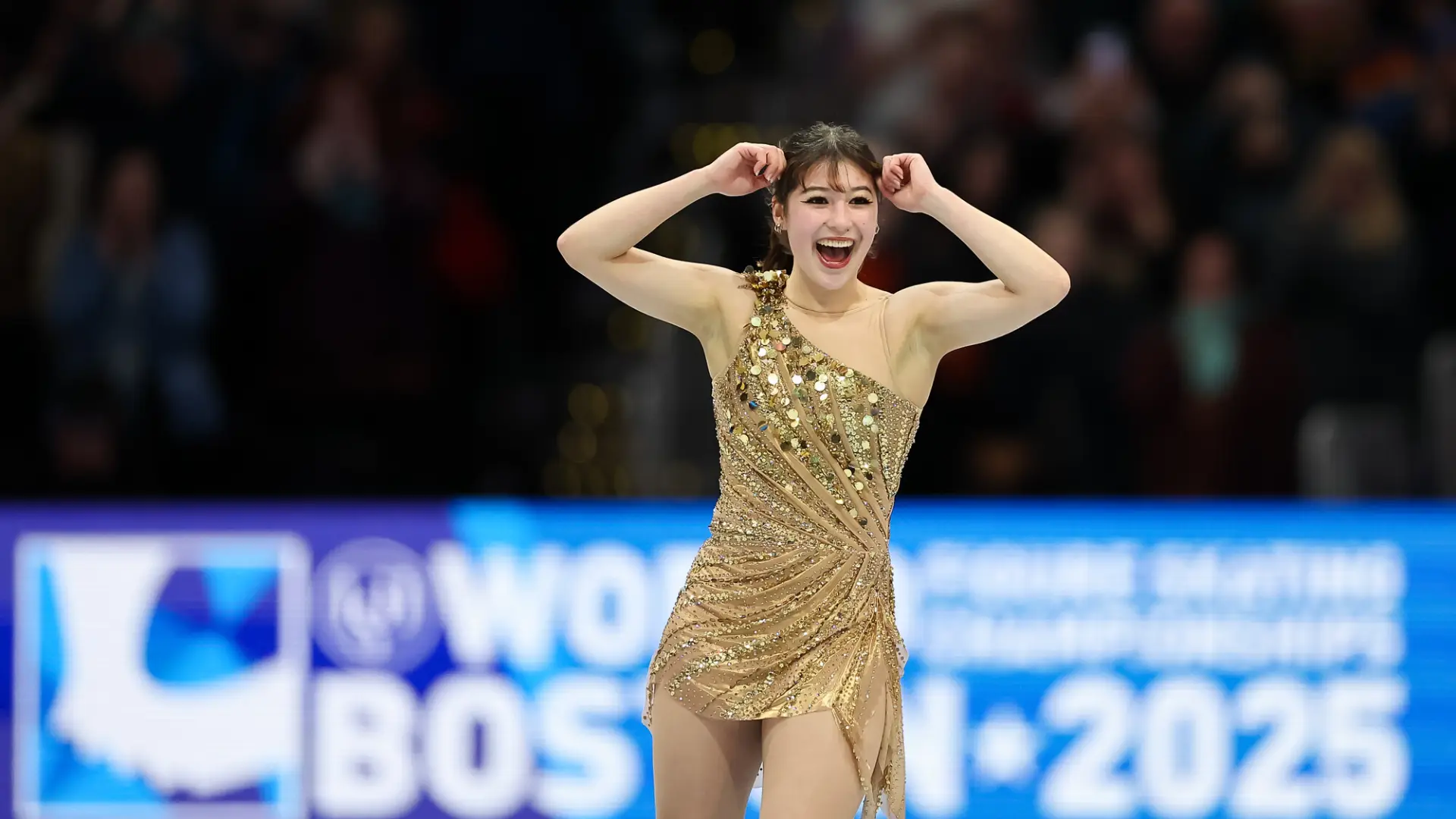 Woman in a gold dress smiles and raises arm in celebration while performing on ice at figure skating competition