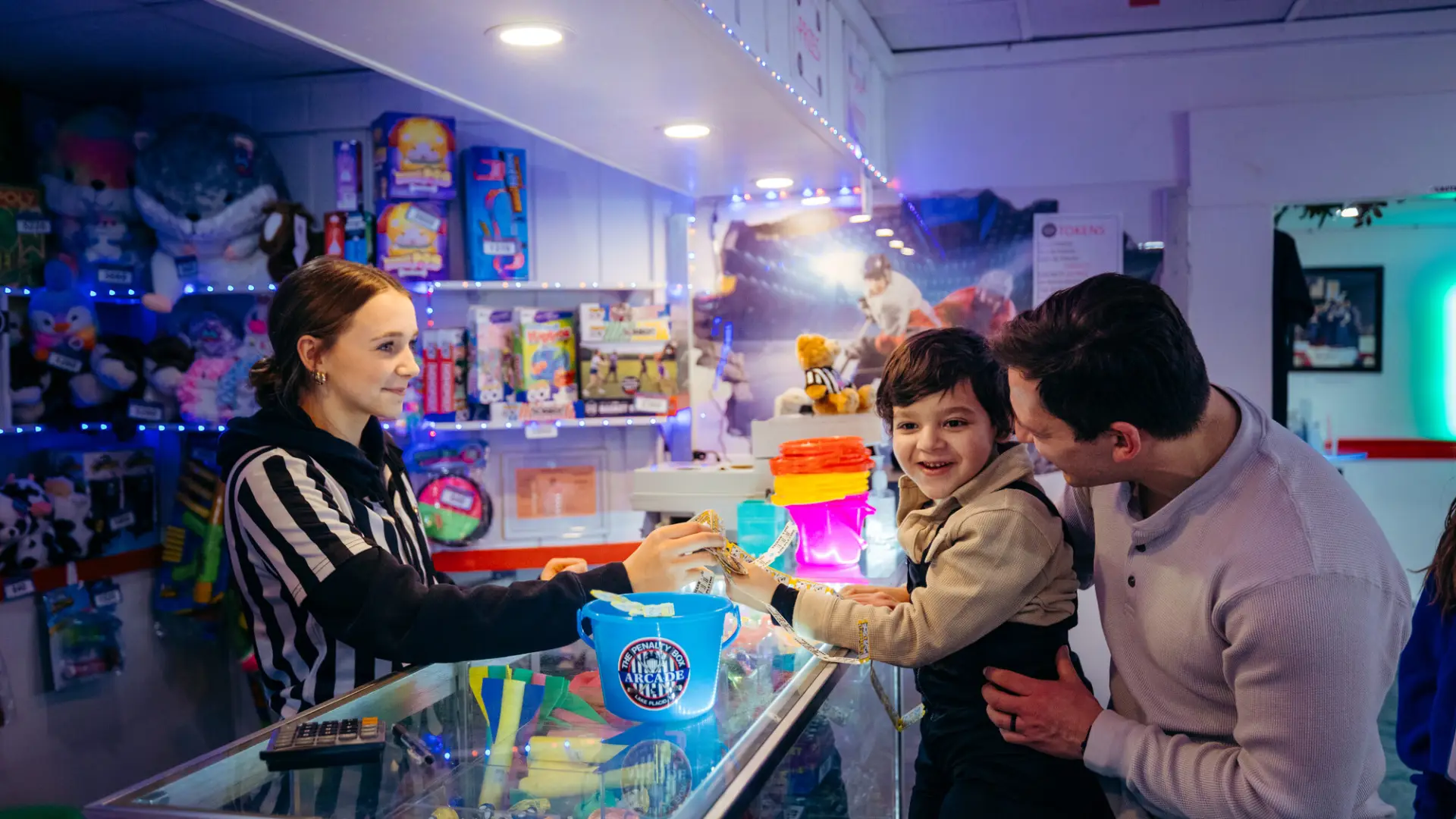 Woman in black and white striped ref-style top hands something across counter to small boy held by father at arcade