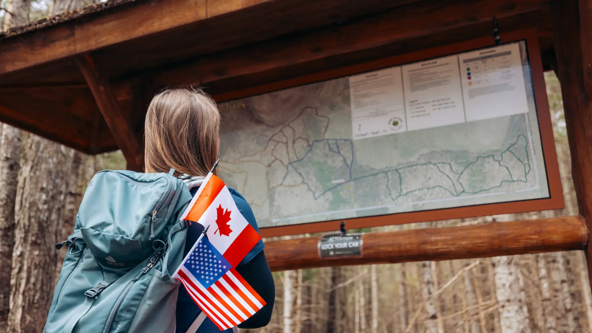 A woman wearing a backpack looks at a hiking kiosk. Small USA and Canadian flags stick up out of the backpack.