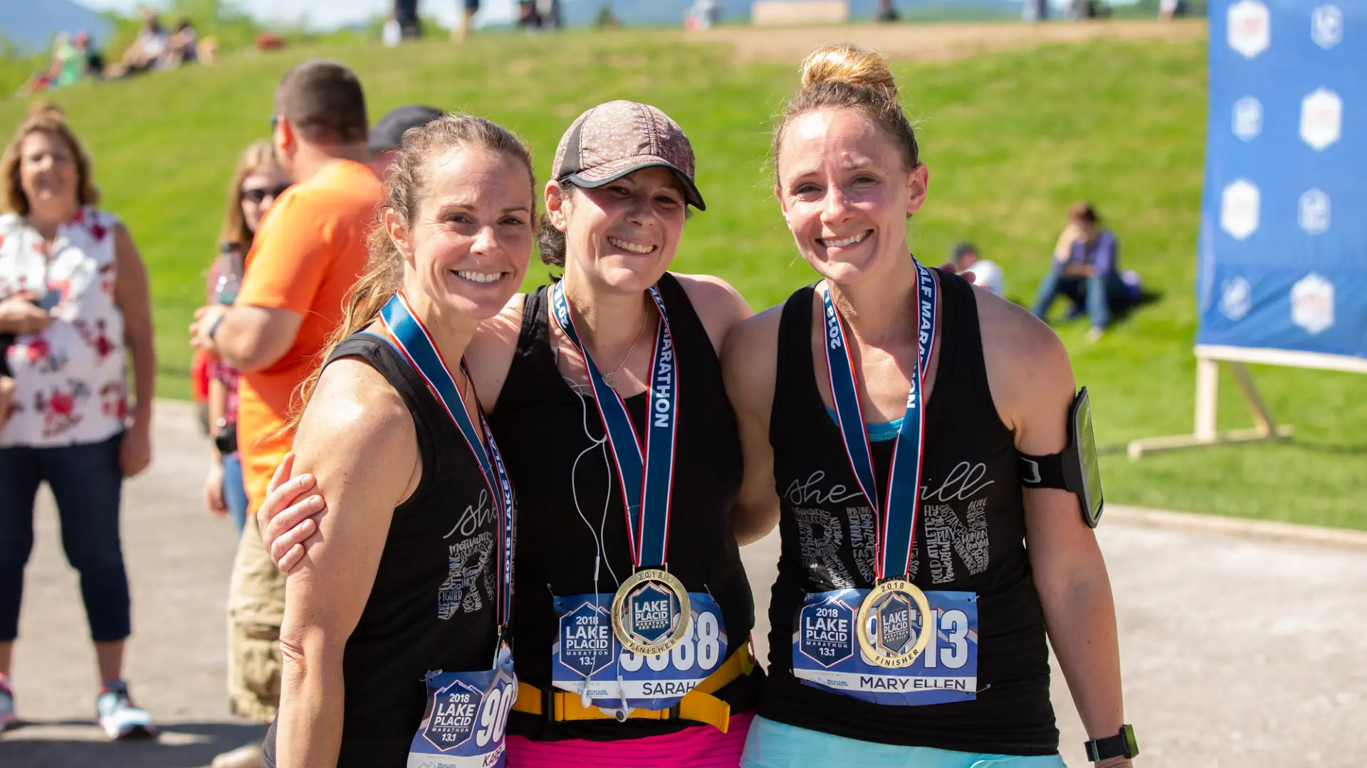 Three women wearing sports clothing and race numbers stand with arms around each other smiling wearing race finisher medals