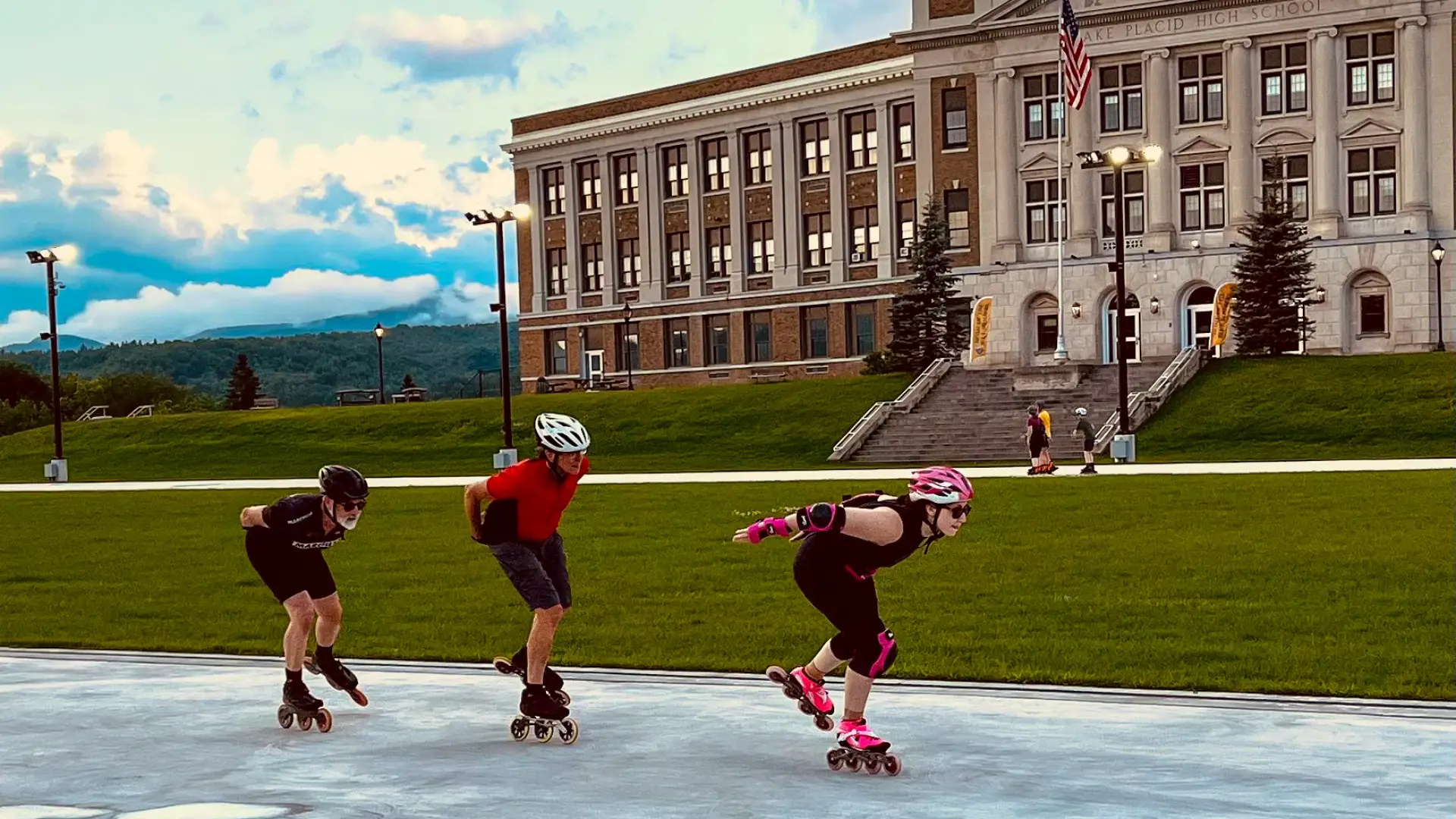 Three adults in helmets and skating clothes inline skate in a row, with other skaters and a school in the background.