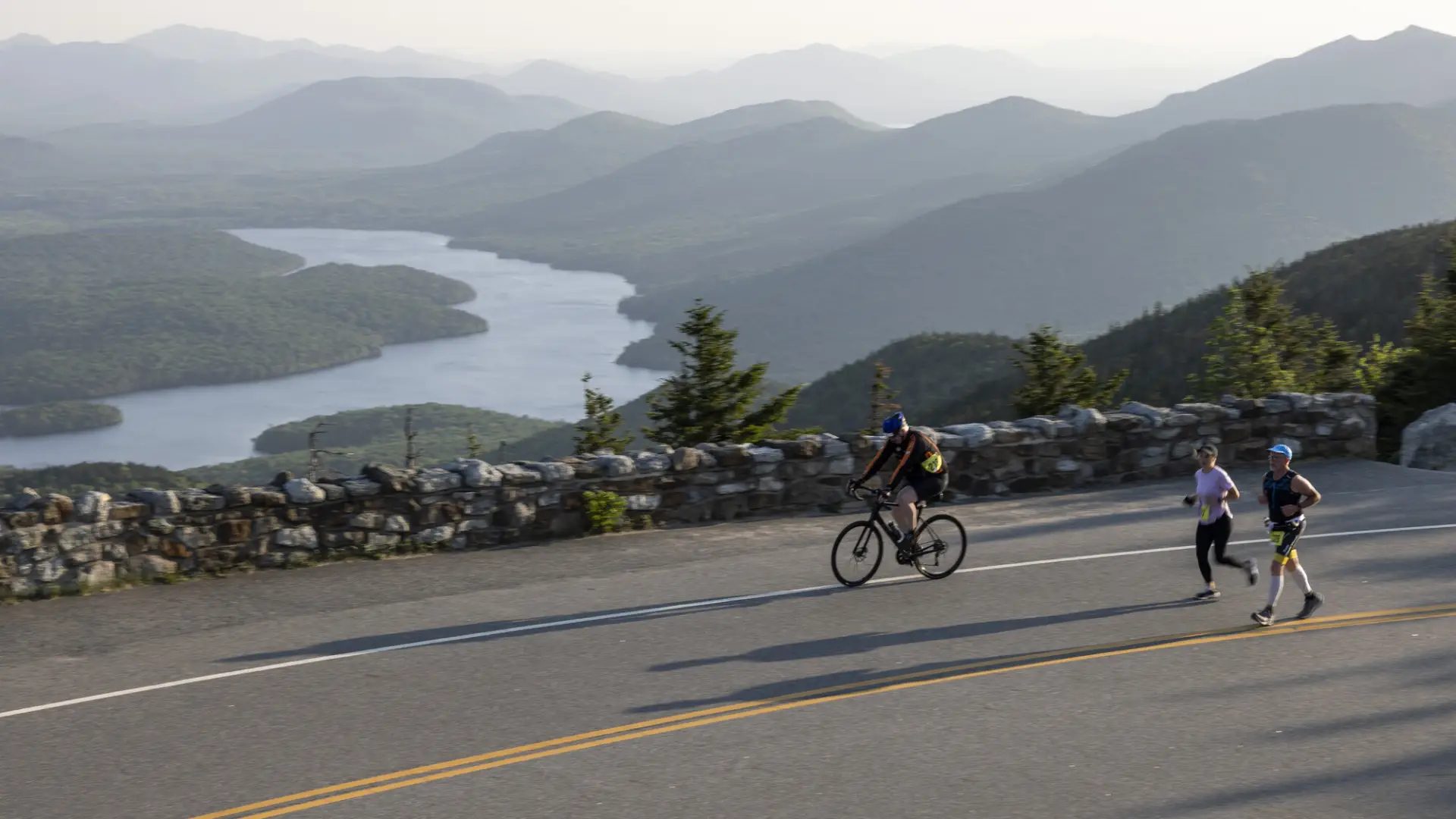 A cyclist and runners going up a road with mountain and lake views