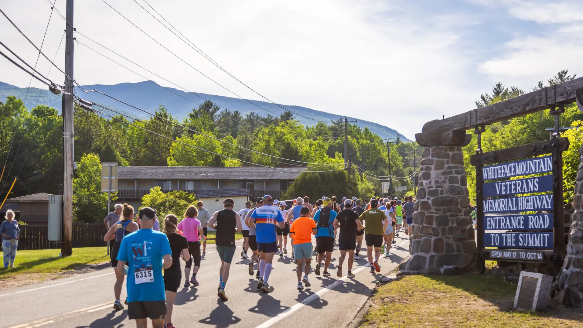 A large group of runners head down a road, passing a stone sign for the Whiteface Mountain Veterans Memorial Highway.