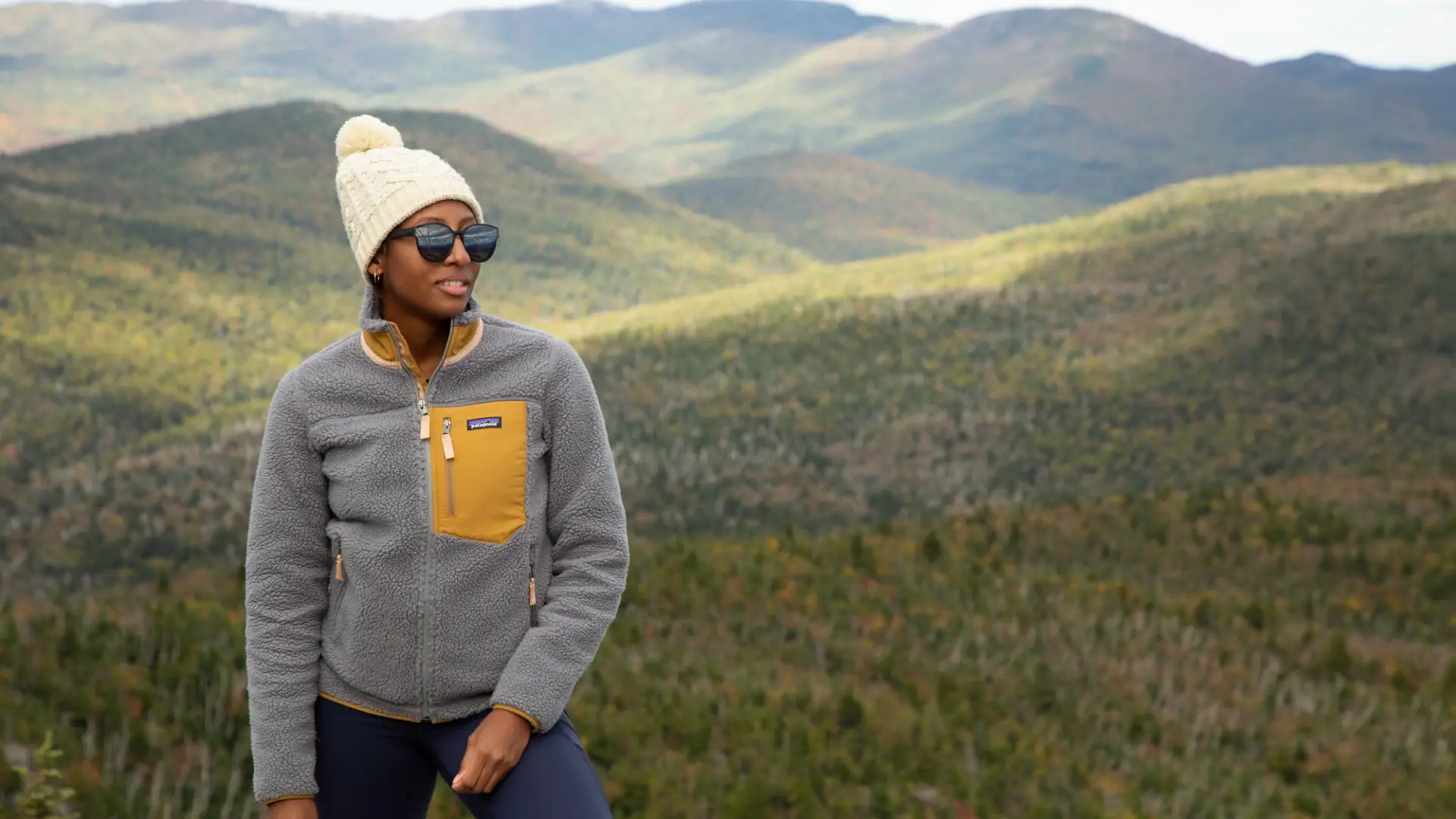 A hiker smiles facing the camera with a mountainous landscape in the background during late fall