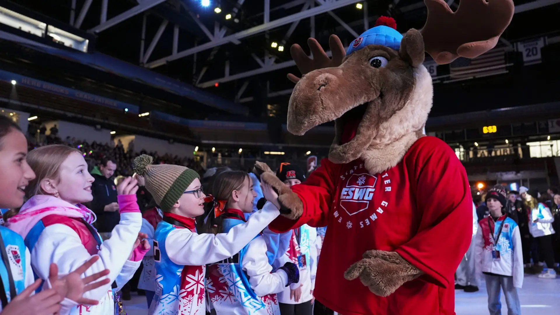 A group of children in colorful winter jackets high-five a person in a cheerful moose mascot costume at an indoor event, conveying excitement and joy.