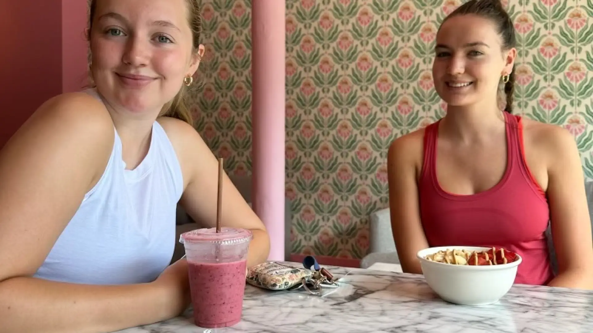 Two young women smile over a light lunch in a small cafe with pink and green decor.