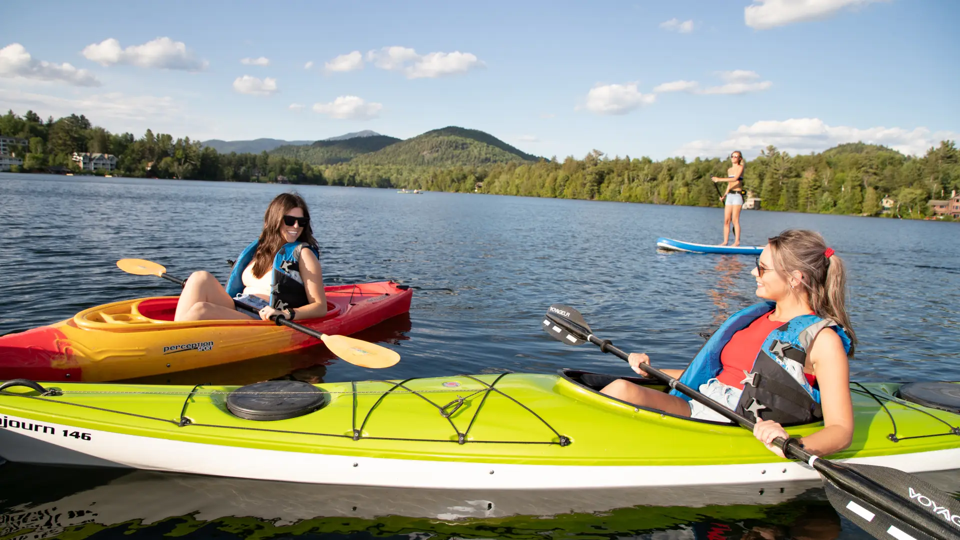 Two women kayak and another paddles a SUP on Mirror Lake, all of which can be found at Lake Placid boat rentals.
