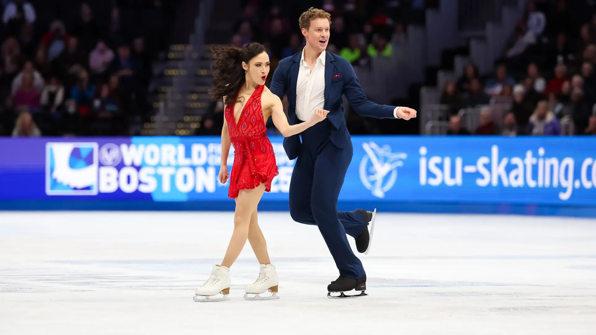 Female figure skater in a red dress and male figure skater in a blue suit perform side by side on ice