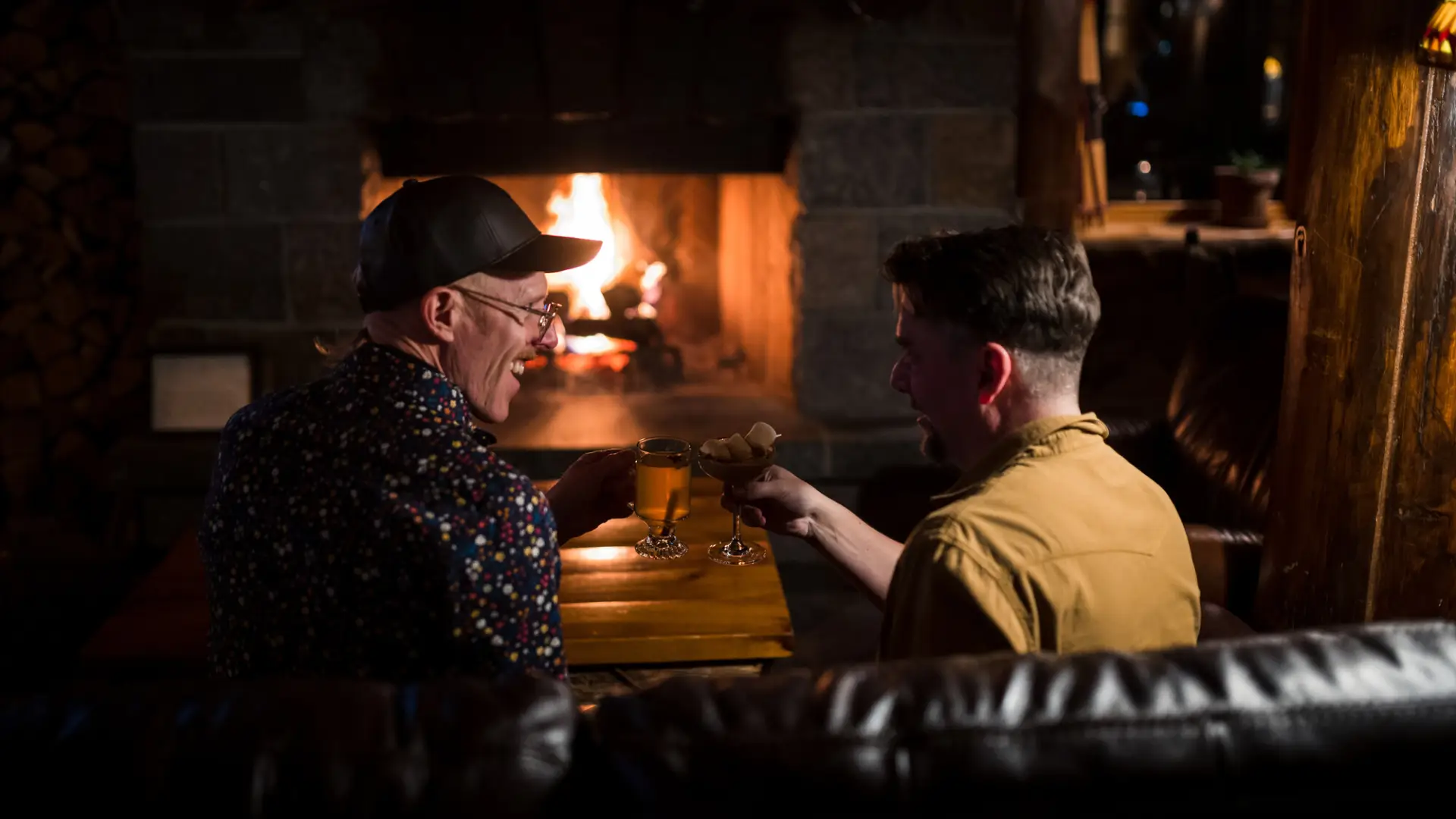 Two men cozy up by the fire with drinks in a dimly lit restaurant.