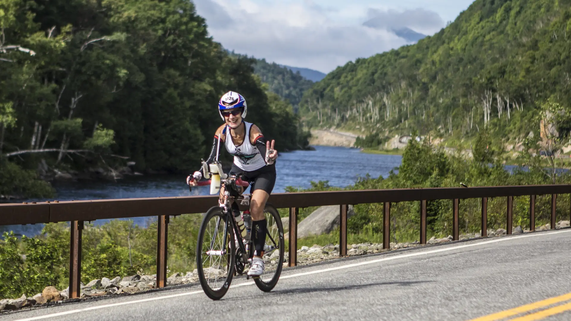 a woman rides her bike and throws up a peace sign in front of a rushing river.