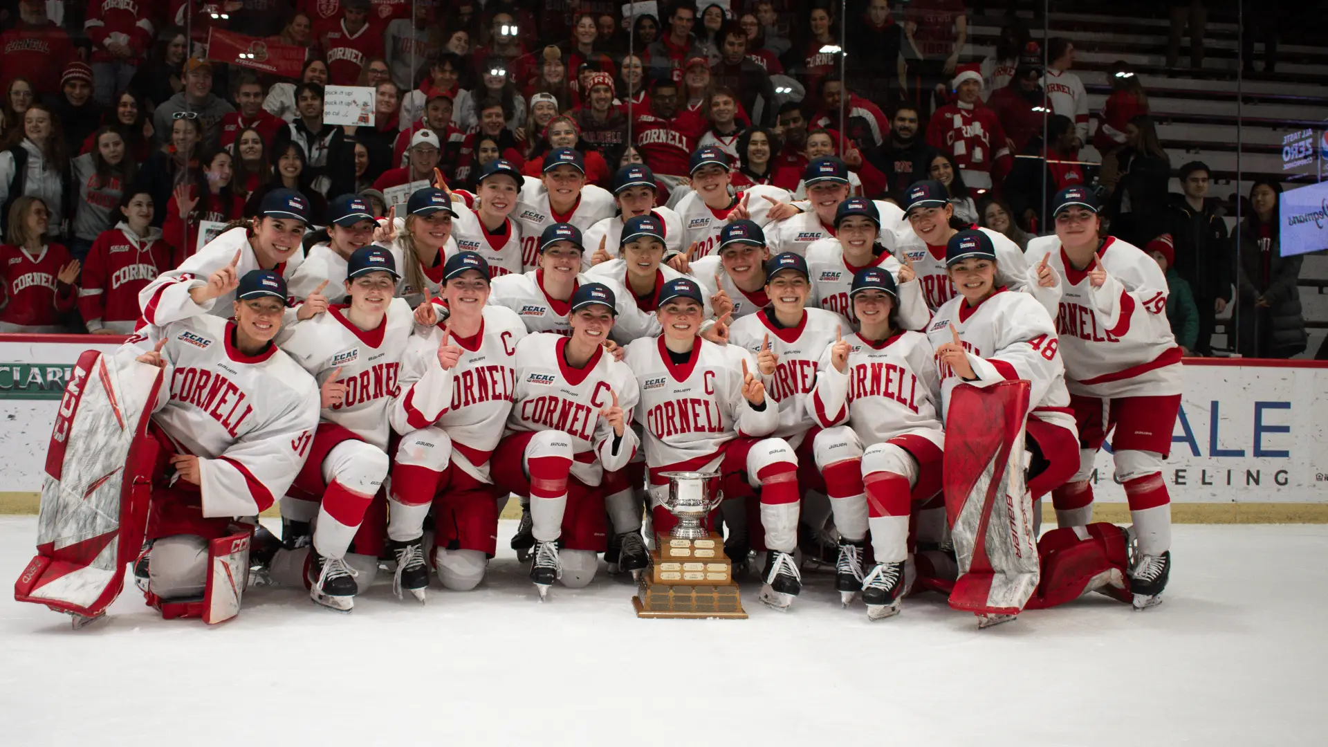 Women's hockey team in uniform and ECAC championship hats gathered for large group photo on ice with trophy