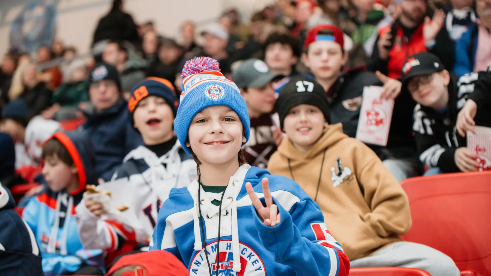 Child in hockey jersey and hat holds up hand in peace sign while sitting in seat of 1980 Herb Brooks Arena along with a crown of people during a hockey game
