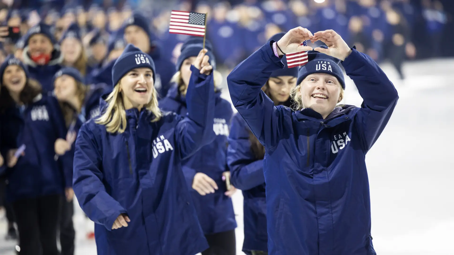 Team USA athletes wearing blue track suit uniforms and beanies walk out on ice during FISU opening ceremony inLake Placid. One girl waves a small American flag while another holds her hands up in the shape of a heart.