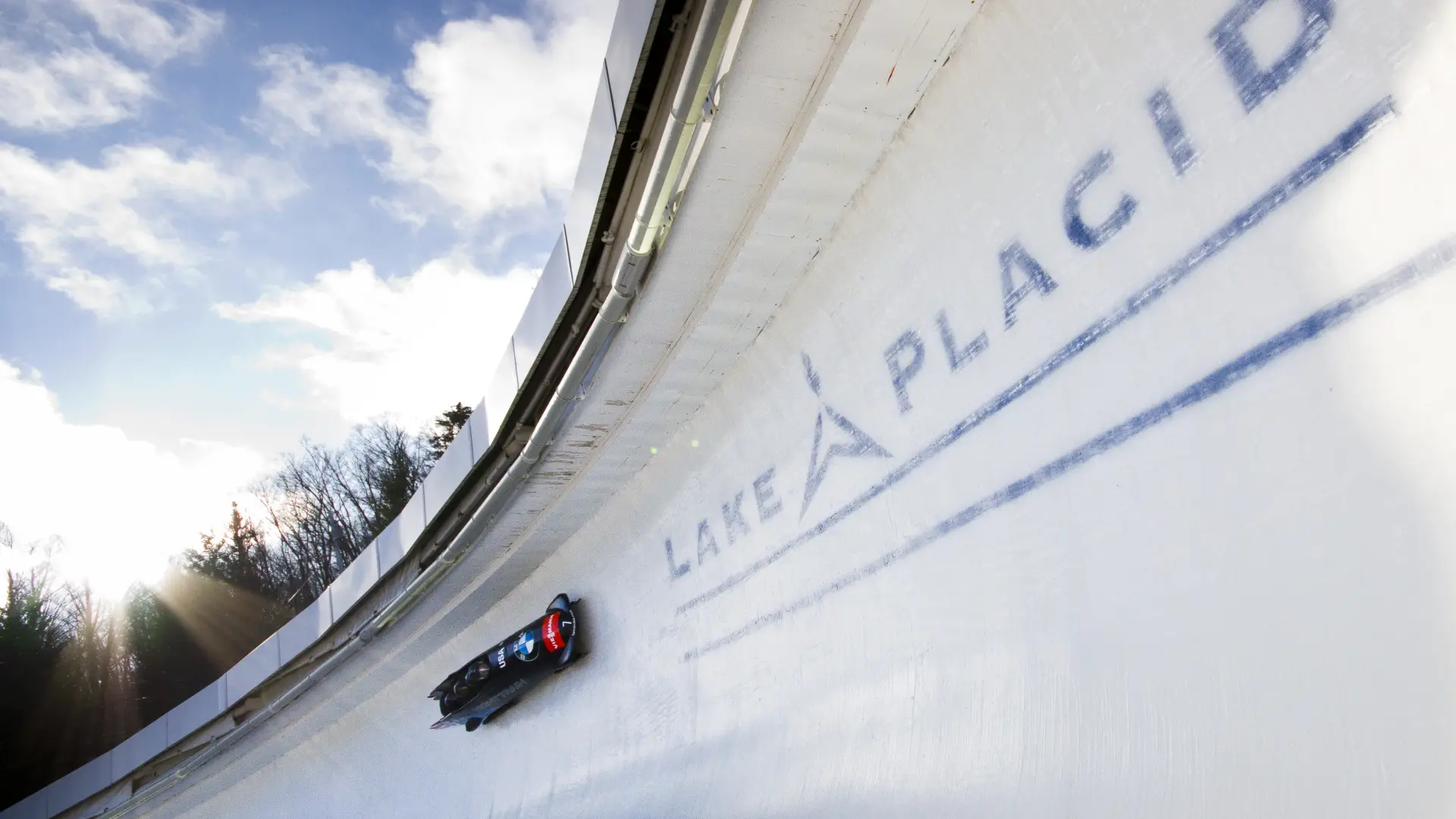 A team takes a large banked curve on the Lake Placid Olympic bobsled track with white clouds and blue sky in the background