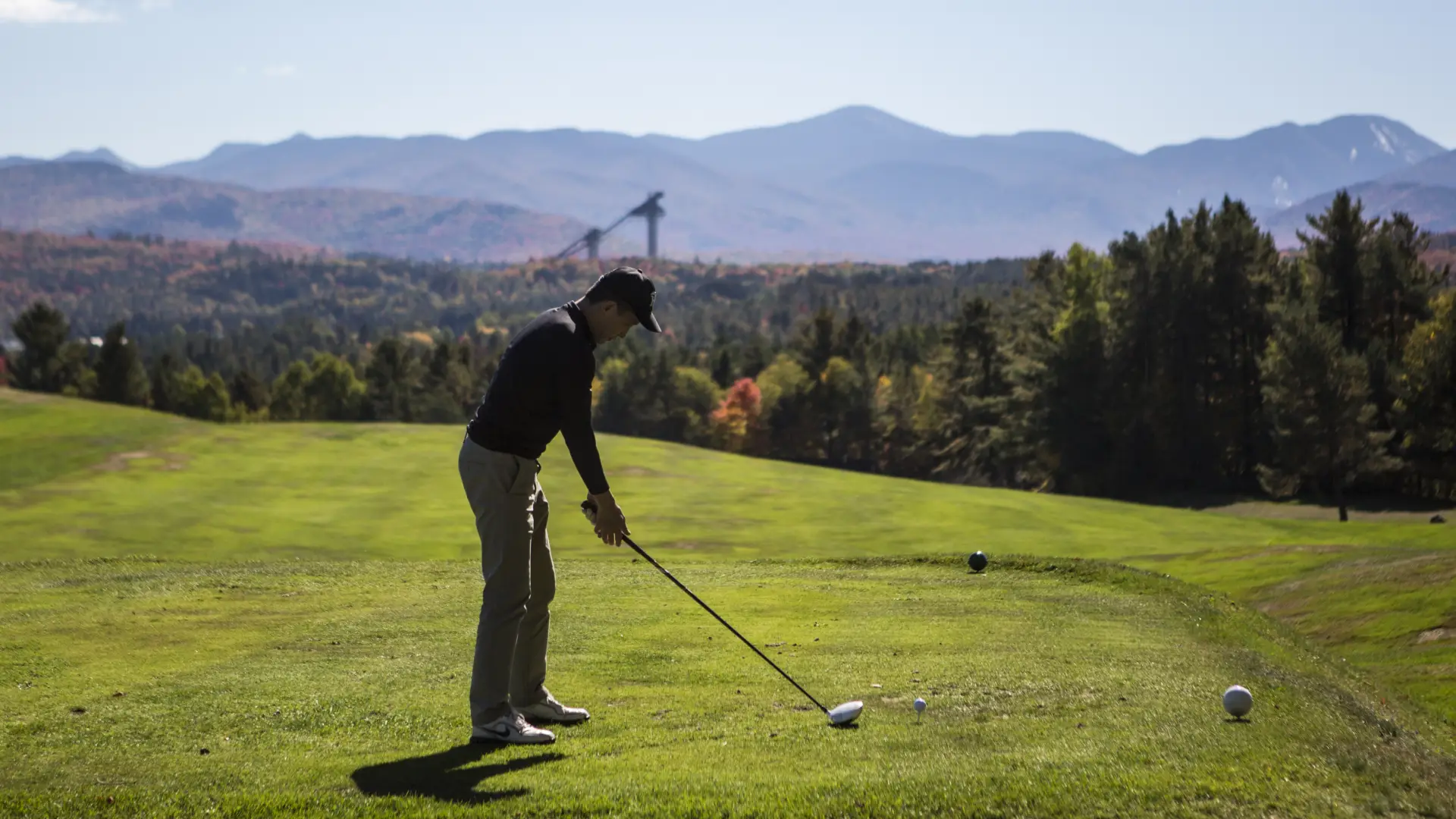 A golfer tees off with the Adirondack High Peaks and Olympic ski jumps in the background.