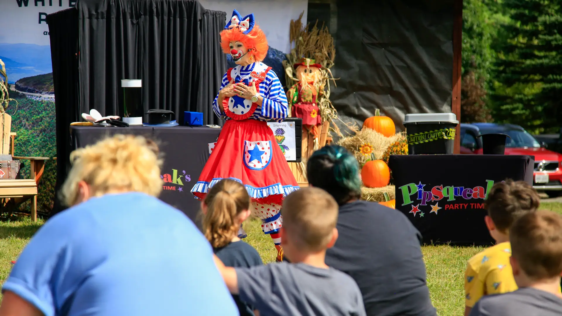 Woman dressed as a clown stands at front of a crowd of children sitting on grass talking to them