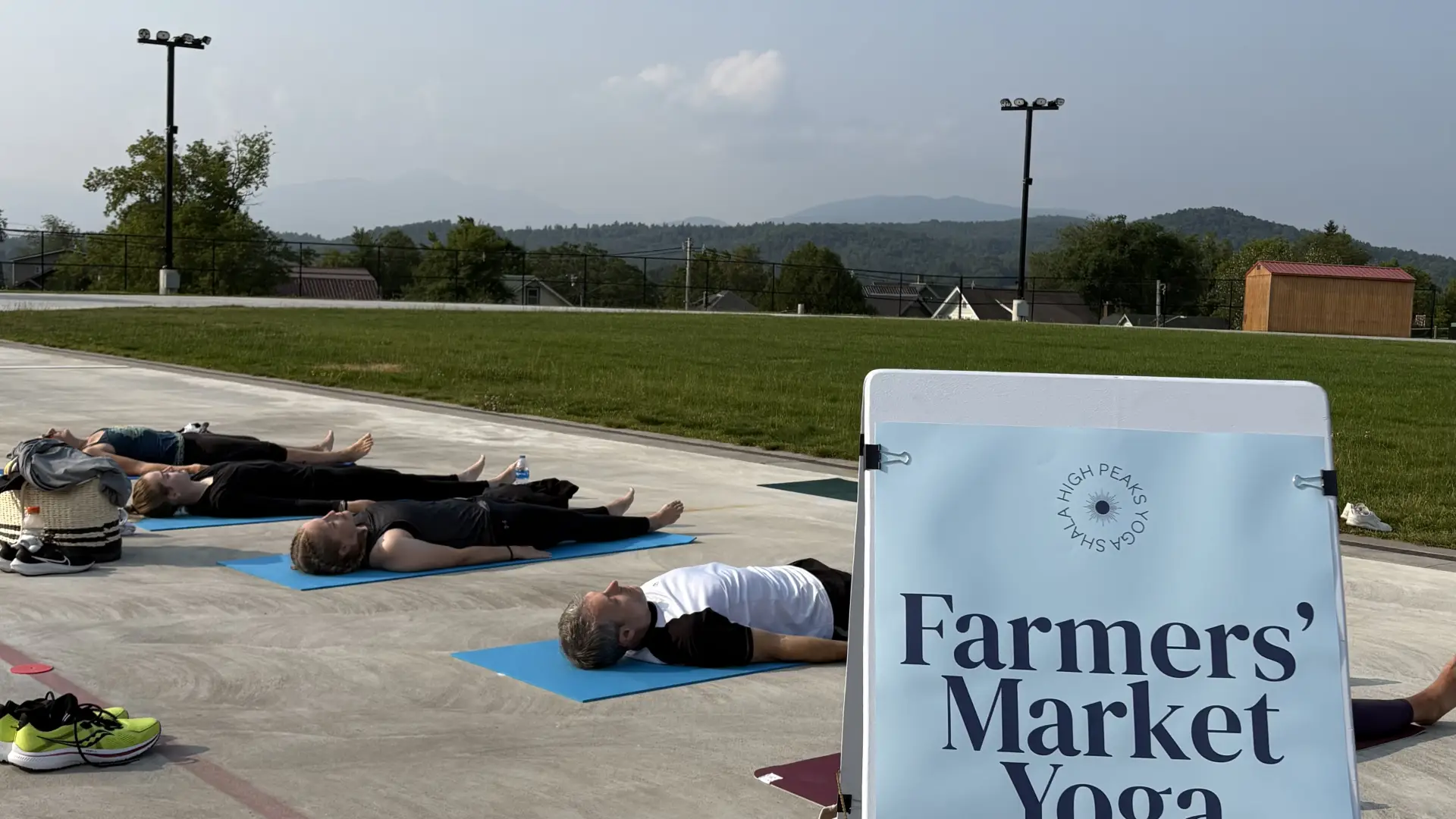 People lay on yoga mats on Olympic Oval track with mountains in distance and banner sign that says Farmers' Market Yoga