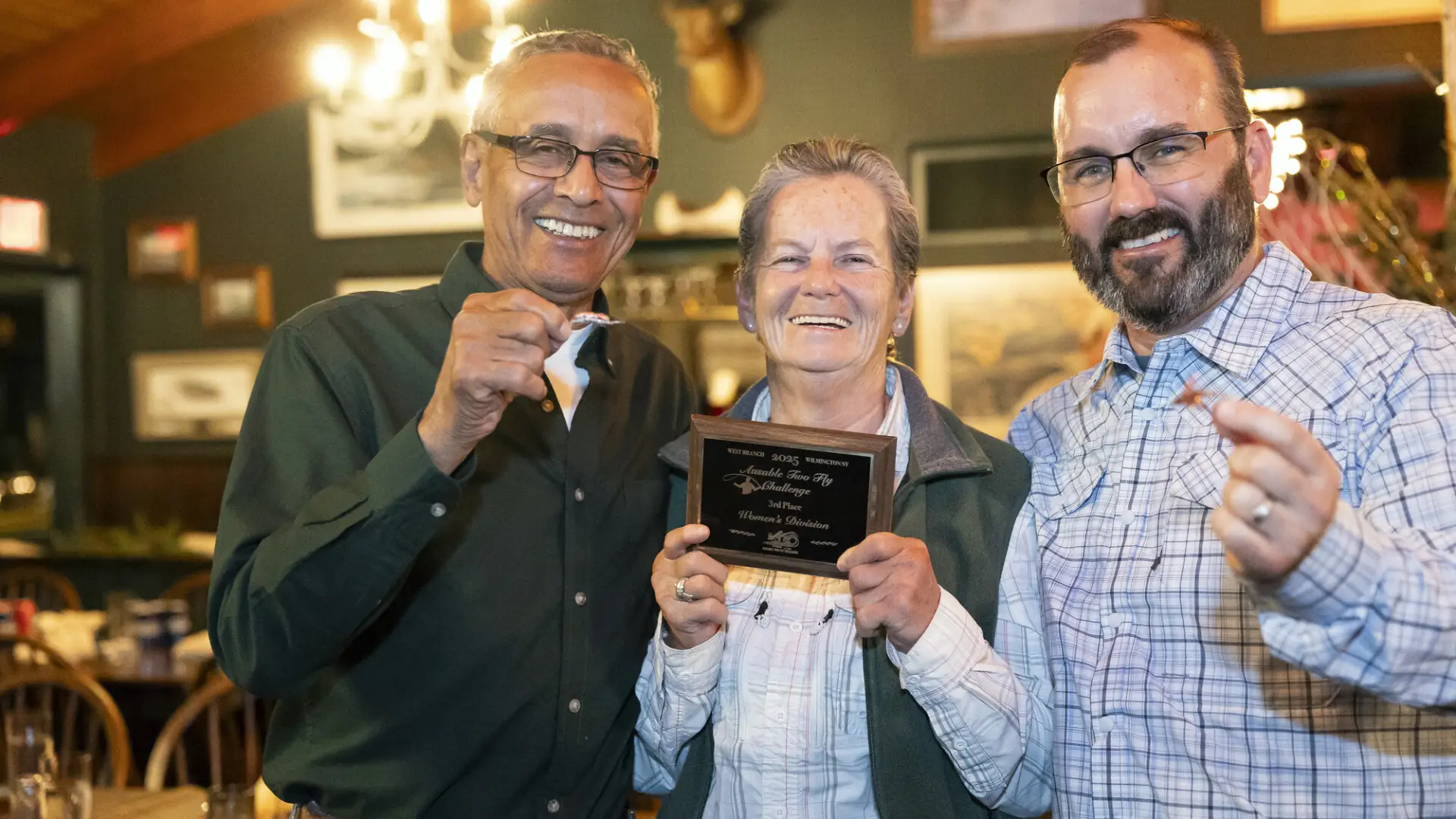 Two men holding up fishing flies stand on either side of a woman holding a plaque at an Ausable River Two-Fly Challenge event