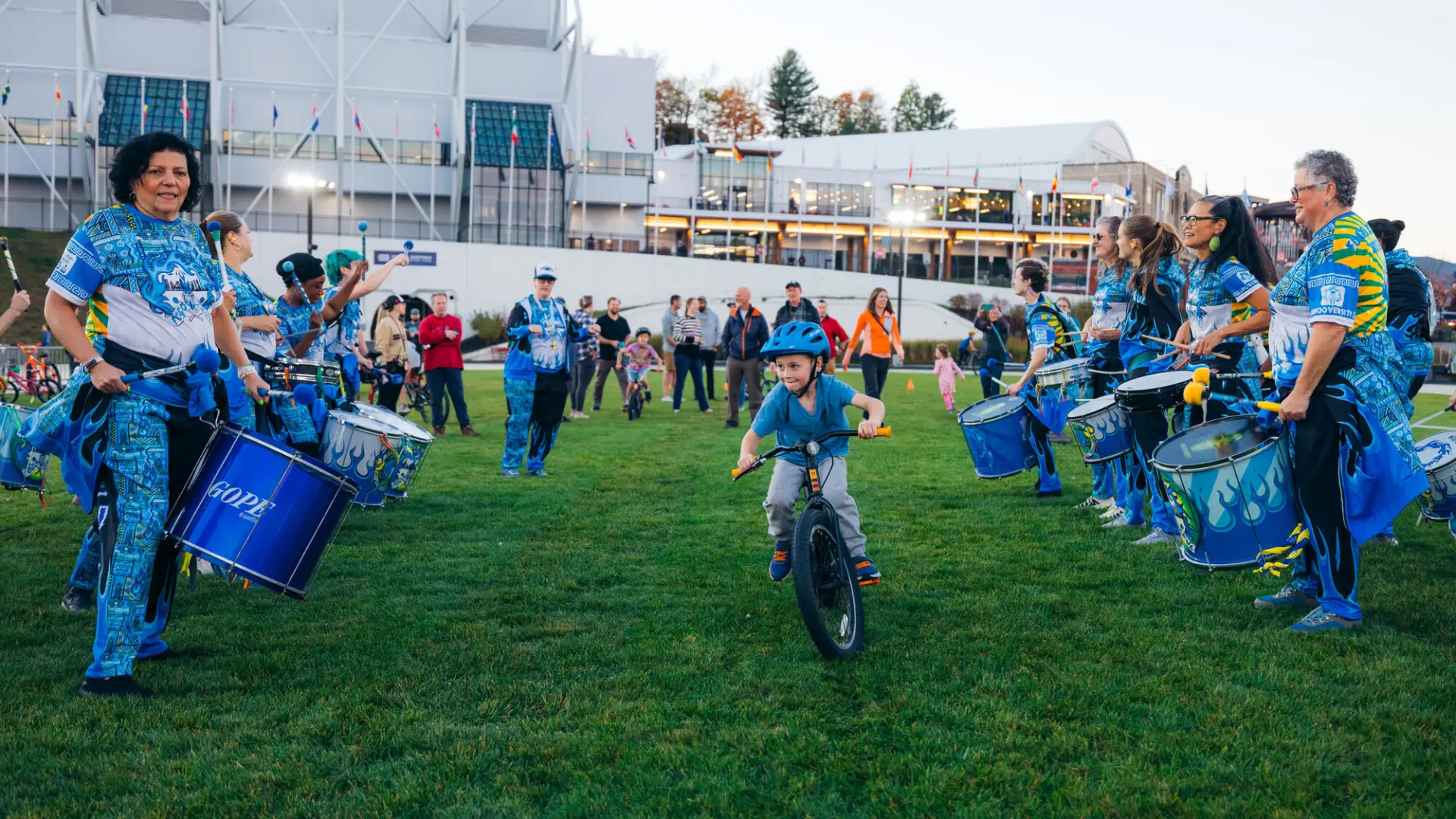 Child rides bike through aisle created by drum line on grass of Olympic Speed Skating Oval