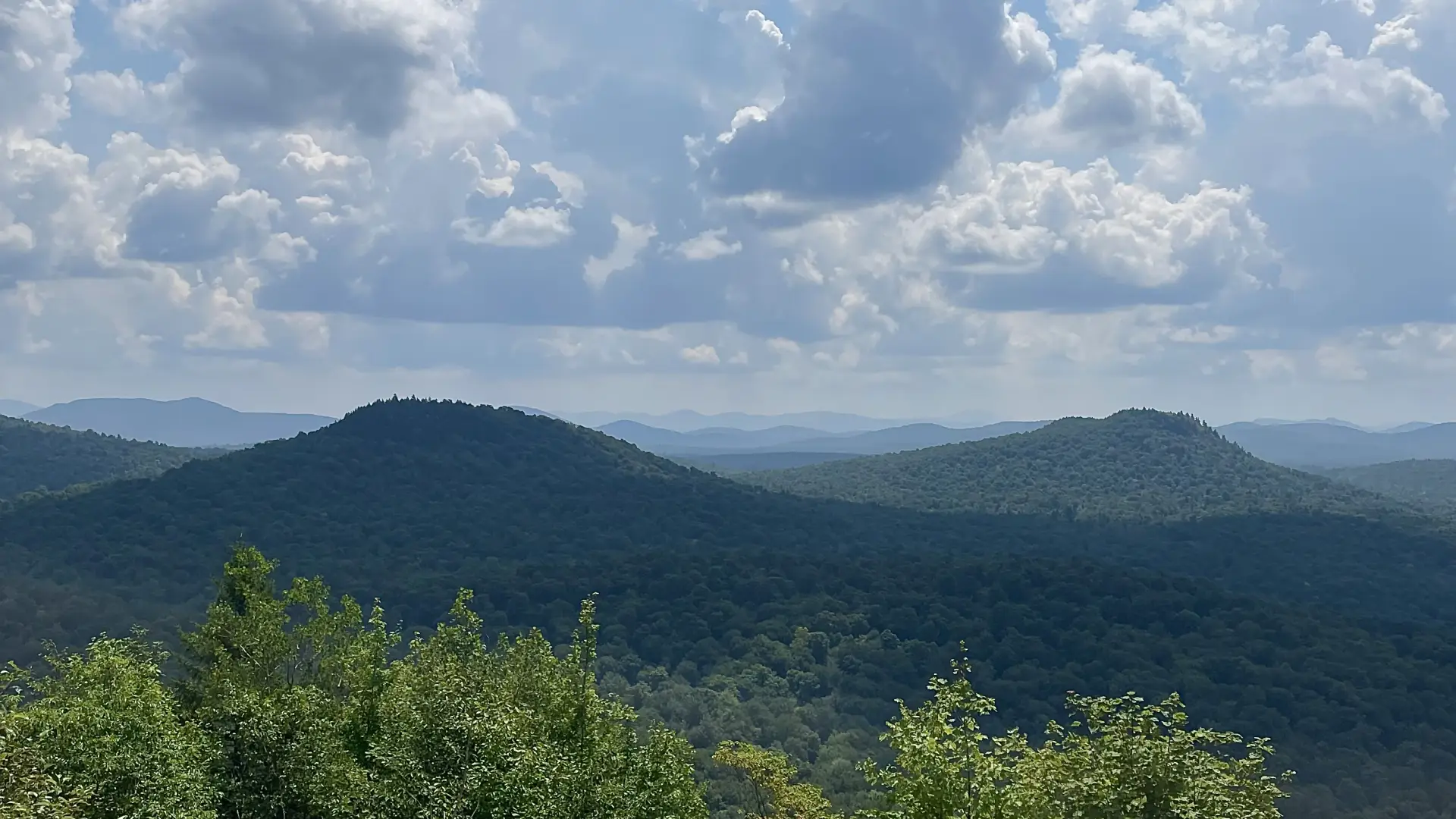Hikers sit and stand on rocky summit of Goodman Mountain looking at and photographing the view.