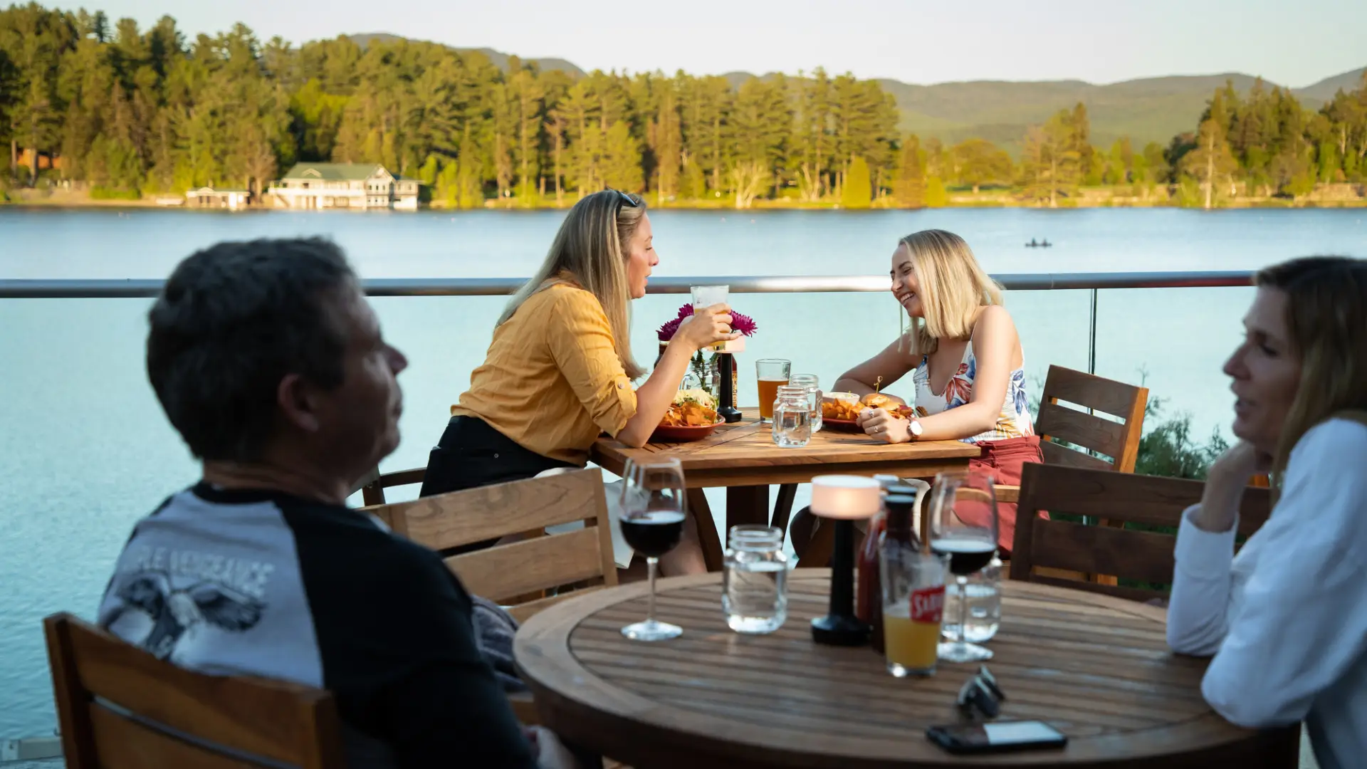 Two women site across from each other at bistro table on deck overlooking lake drinking cocktails with other diners in the foreground