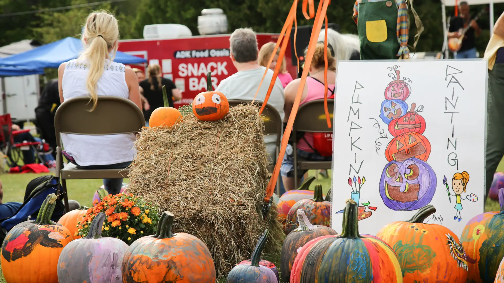 Collection of painted pumpkins and hay bales next to a tent pole and paint a pumpkin sign